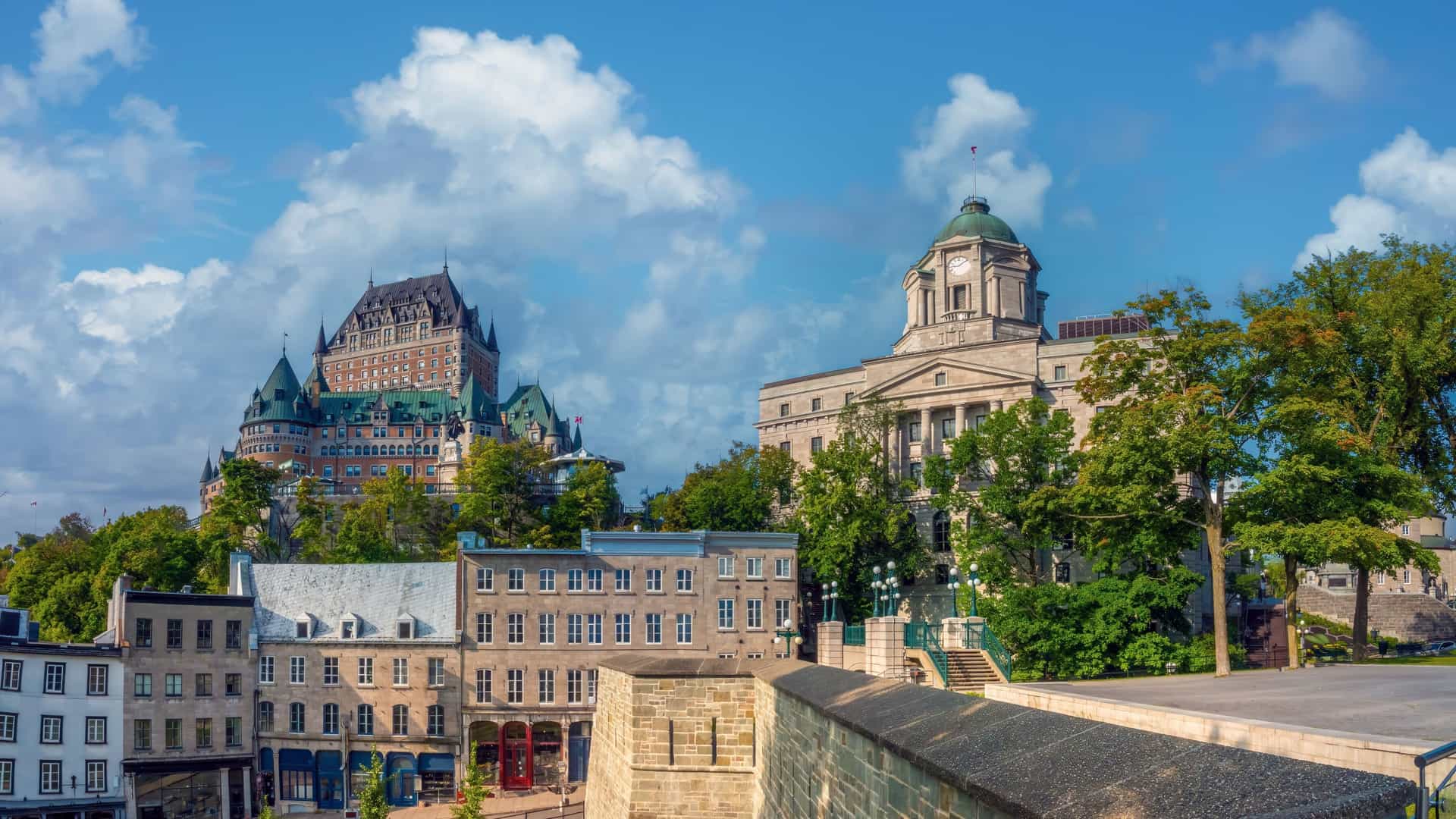 A panoramic view of the Quebec City skyline, highlighting the historic buildings of the Petit Champlain district in the Lower Town, which are clustered below the cliff face, with the iconic Fairmont Le Château Frontenac hotel dominating the Upper Town above.