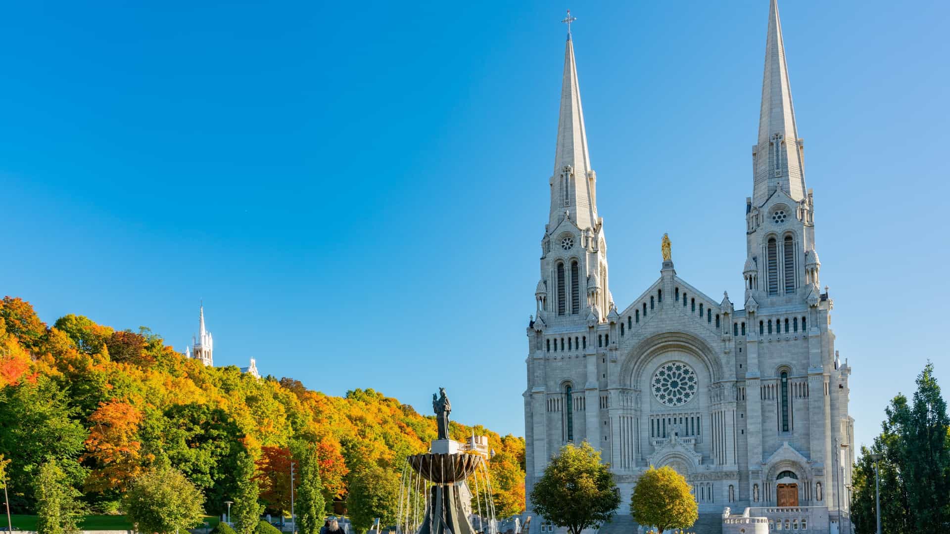 An exterior morning view of the monumental, Neo Romanesque Basilica of Sainte Anne de Beaupré, a Catholic national shrine in Quebec, Canada, with a bright red maple tree visible on the grounds, suggesting an autumn setting.