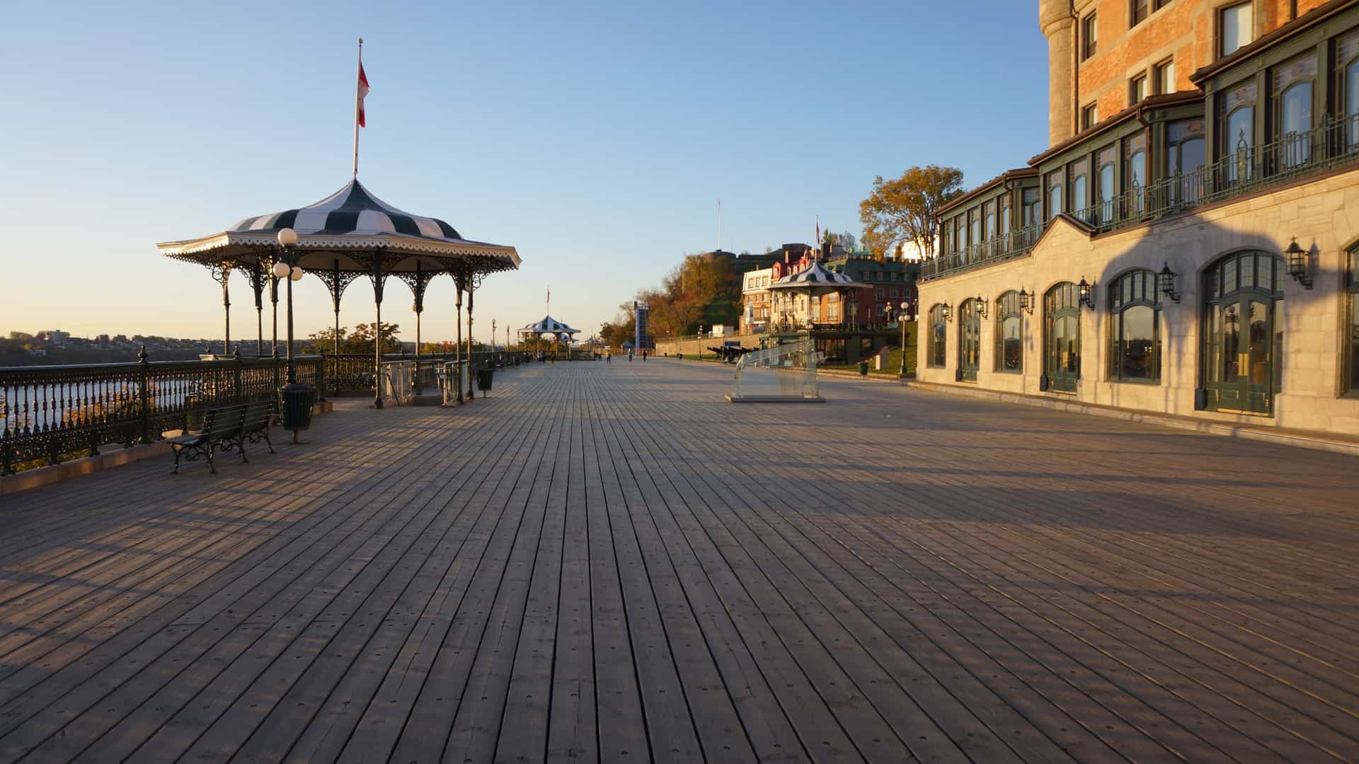 A bright morning view of the Dufferin Terrace promenade in Upper Town, Quebec City, Canada. The wooden walkway, lined with historic cast-iron streetlights and ornate gazebos, leads toward the massive, turreted Fairmont Le Château Frontenac hotel dominating the background. The scene, captured in October, offers a famous view of the city's UNESCO World Heritage site architecture and the Saint Lawrence River.