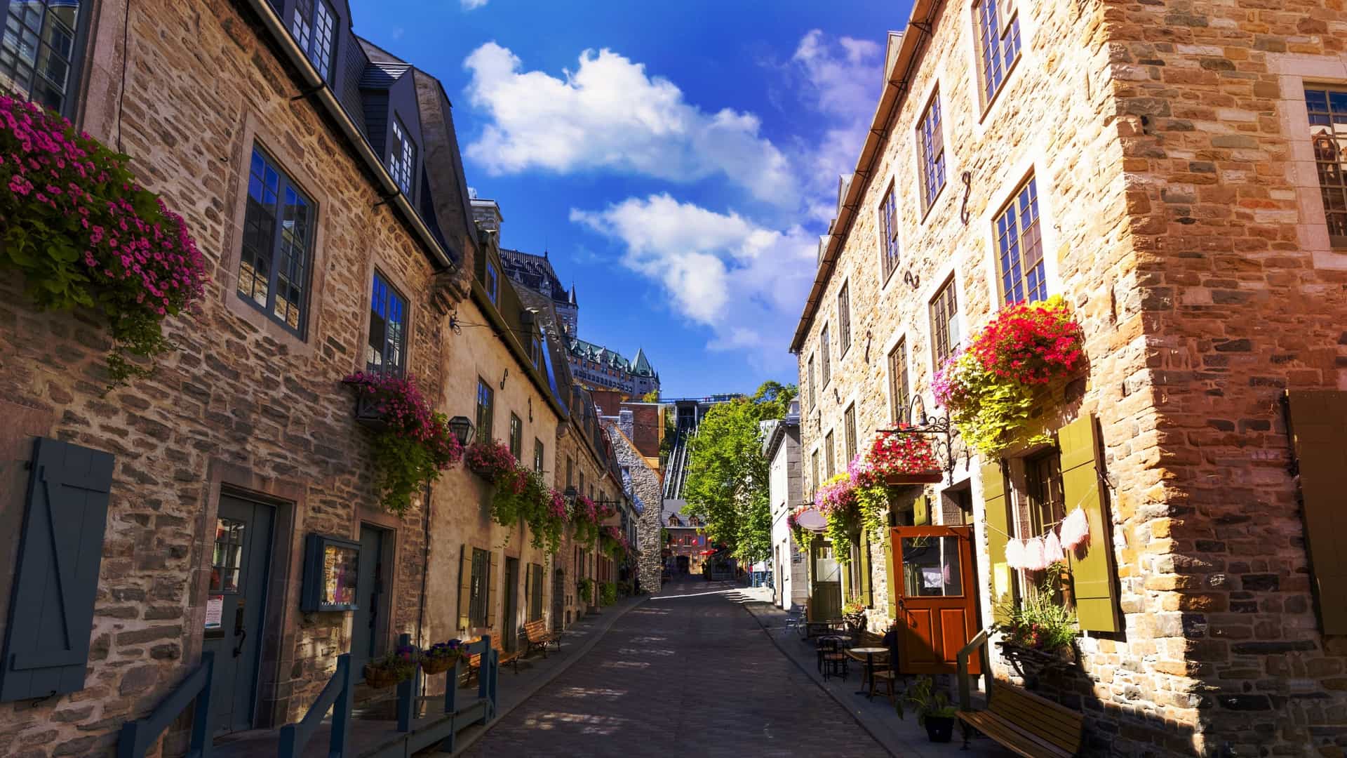 A charming street view of Old Quebec City, Canada, particularly the lower town area. The cobblestone street is lined with quaint, historic stone buildings featuring colorful window boxes and shutters. A vintage-style streetlamp hangs over the narrow road, and a few pedestrians are visible, capturing the European ambiance of the UNESCO World Heritage site.