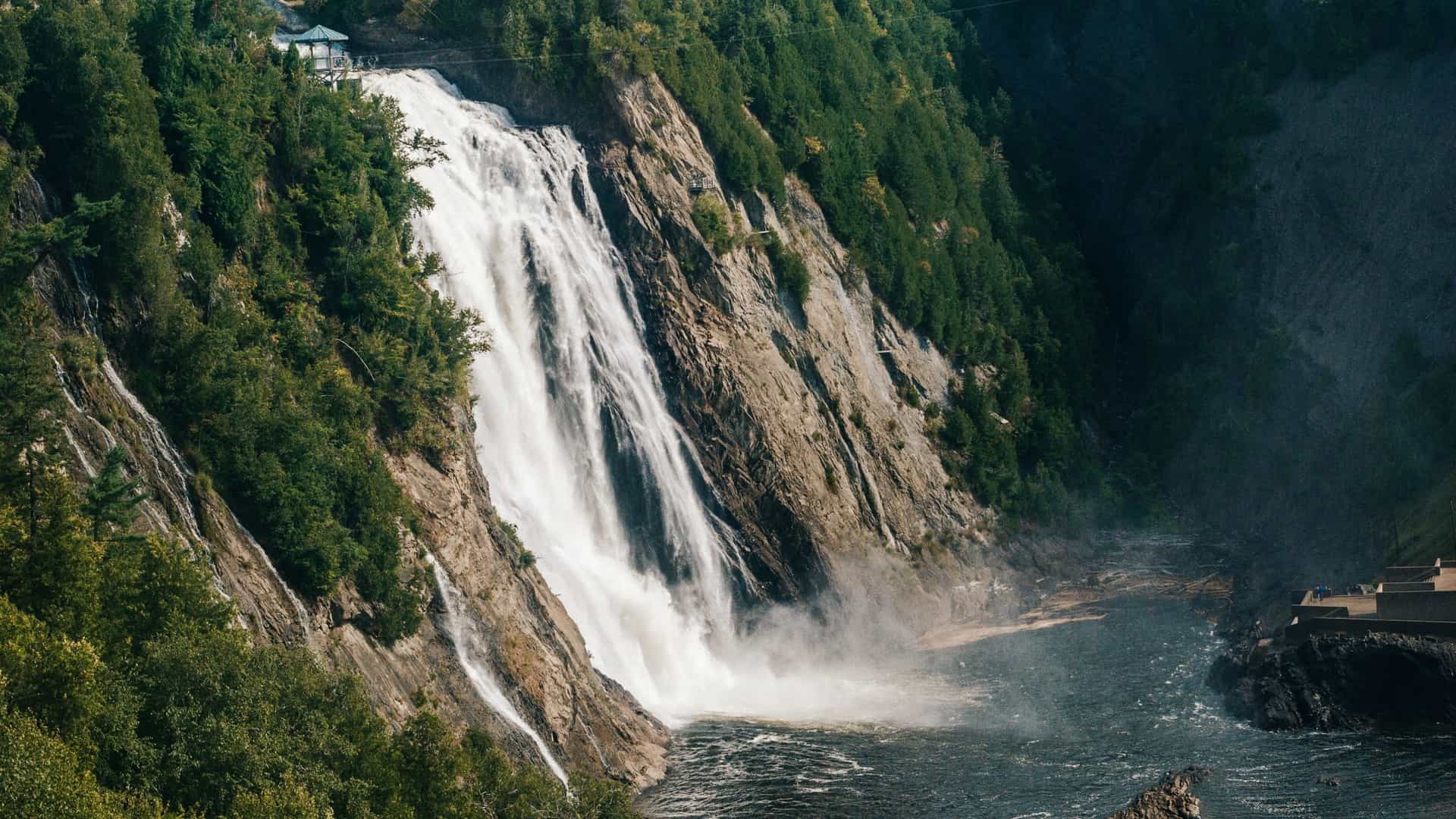A dramatic photograph of the powerful Montmorency Falls (Chute-Montmorency) near Quebec City, Canada. A massive volume of white water plunges down a steep, rocky cliff face into the Montmorency River below. Viewing structures and a bridge are visible near the top of the waterfall, which is framed by lush green forest and steep, dark cliffs.