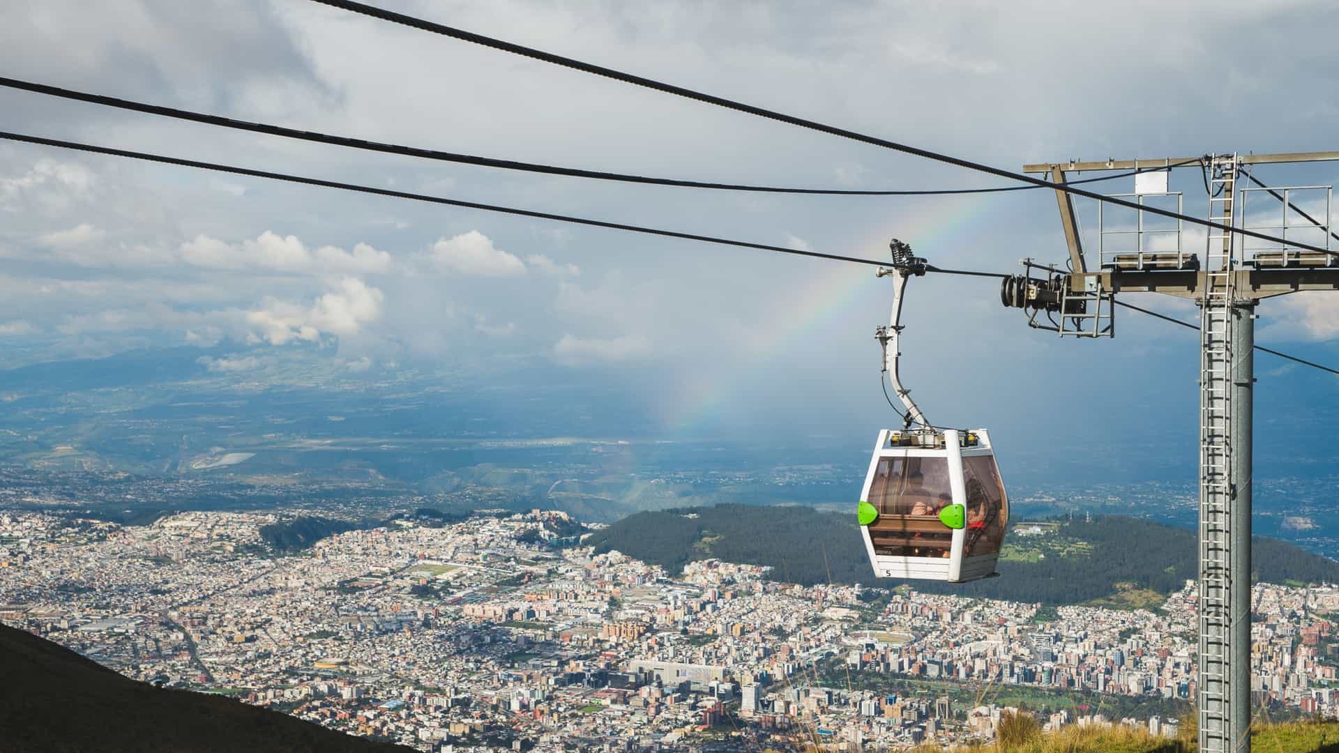 A dramatic high-angle photograph of the TelefériQo cable car in Quito, Ecuador. A white gondola cabin is suspended on the line, passing a support tower, with a rainbow visible against the cloudy sky. In the background, the vast, densely built urban landscape of Quito spreads across the valley floor beneath the slopes of the Pichincha Volcano.