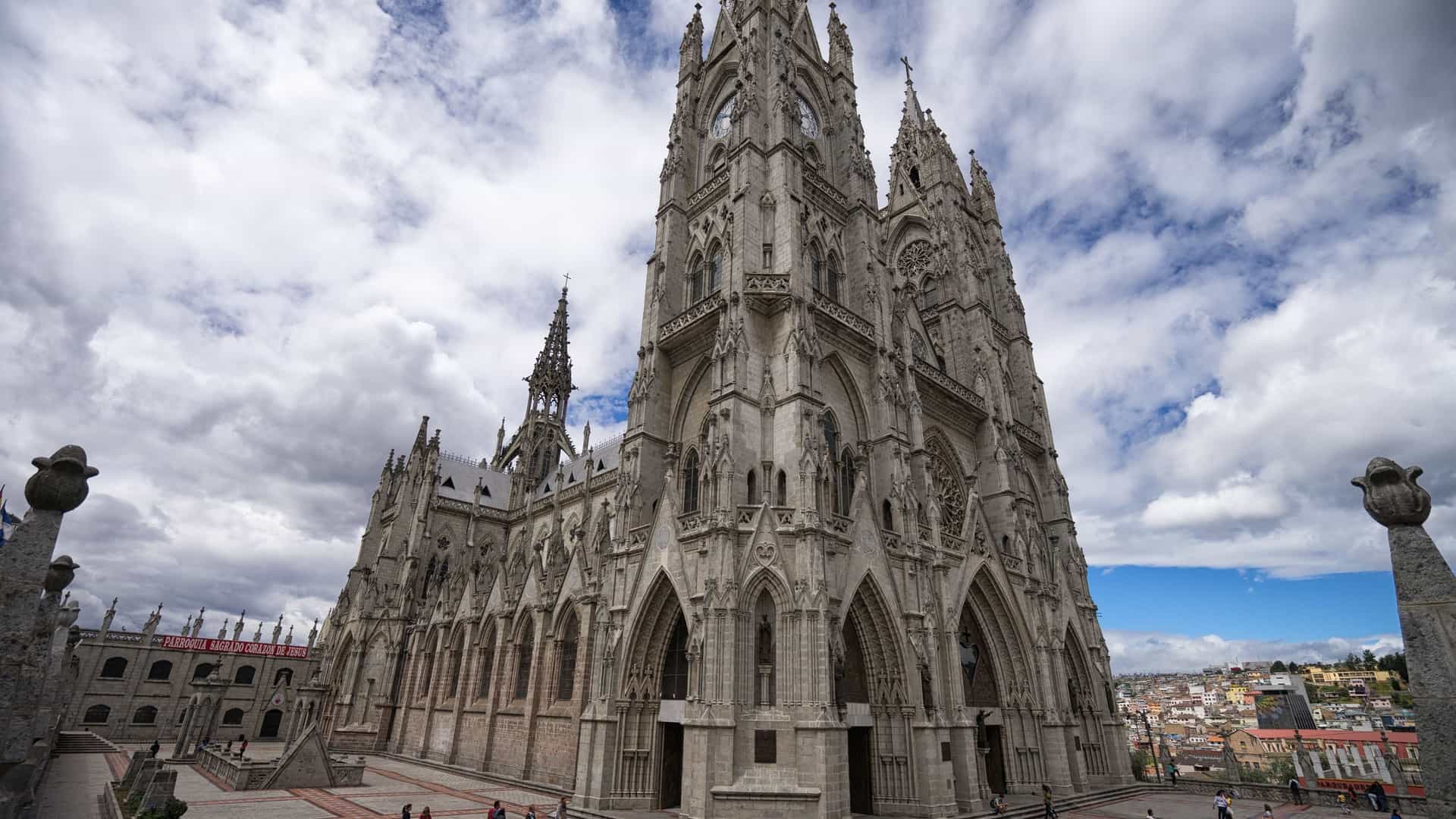A close-up view of the massive Basílica del Voto Nacional in Quito, Ecuador, the largest Neo-Gothic basilica in the Americas. The image highlights the intricate details of its stone facade, featuring tall, pointed arches and its twin soaring towers/spires. Sculpted figures of native Ecuadorian animals (such as iguanas or turtles) are visible, replacing traditional gargoyles.