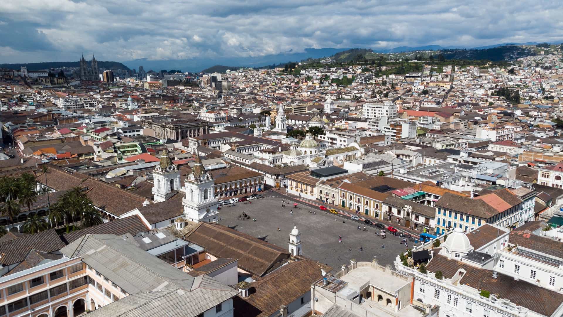 An aerial panoramic view of the Historic Center of Quito, Ecuador. The image displays the dense, sprawling grid of the colonial city with its characteristic terracotta rooftops and narrow streets, set within the mountainous Andean landscape. In the mid-ground, the iconic El Panecillo hill rises, crowned by the towering, winged Virgen de Quito (Virgin of Quito) statue, which overlooks the capital city.