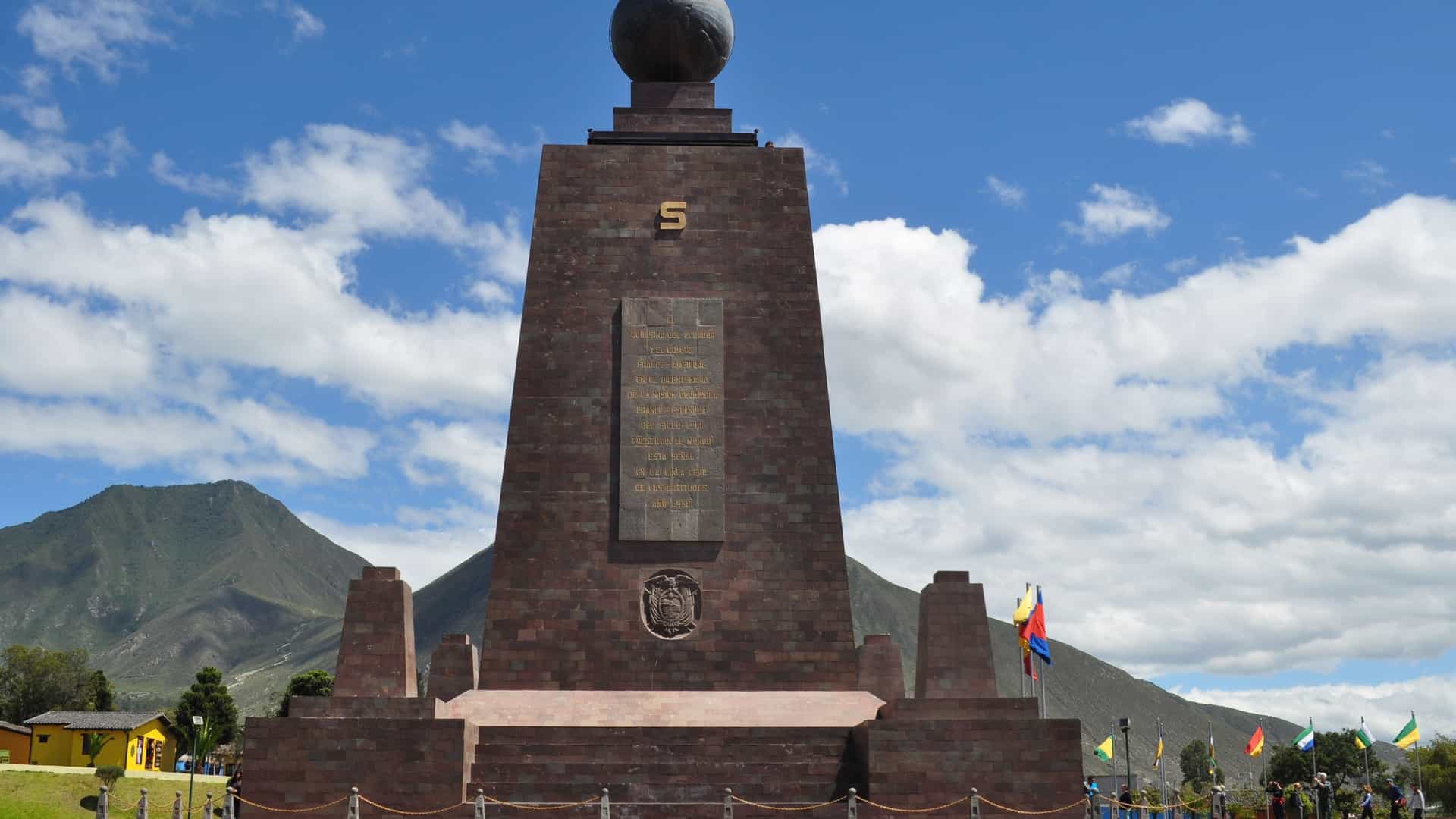 A photograph of the Monumento a la Mitad del Mundo (Middle of the World Monument) located near Quito, Ecuador. The monument is a tall, pyramidal structure topped with a large brass globe. A bright yellow line is painted on the ground and runs up the monument, symbolically marking the equatorial line, where visitors often stand with one foot in the Northern Hemisphere and one in the Southern Hemisphere.