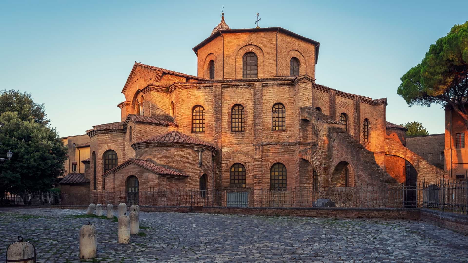 A view inside the Basilica di San Vitale in Ravenna, Italy, showcasing the magnificent 6th century Byzantine mosaics of the presbytery and apse. The surfaces are covered in dazzling gold, green, and blue tesserae, with intricate patterns and figurative scenes, leading up to the vaulted ceiling and arches.