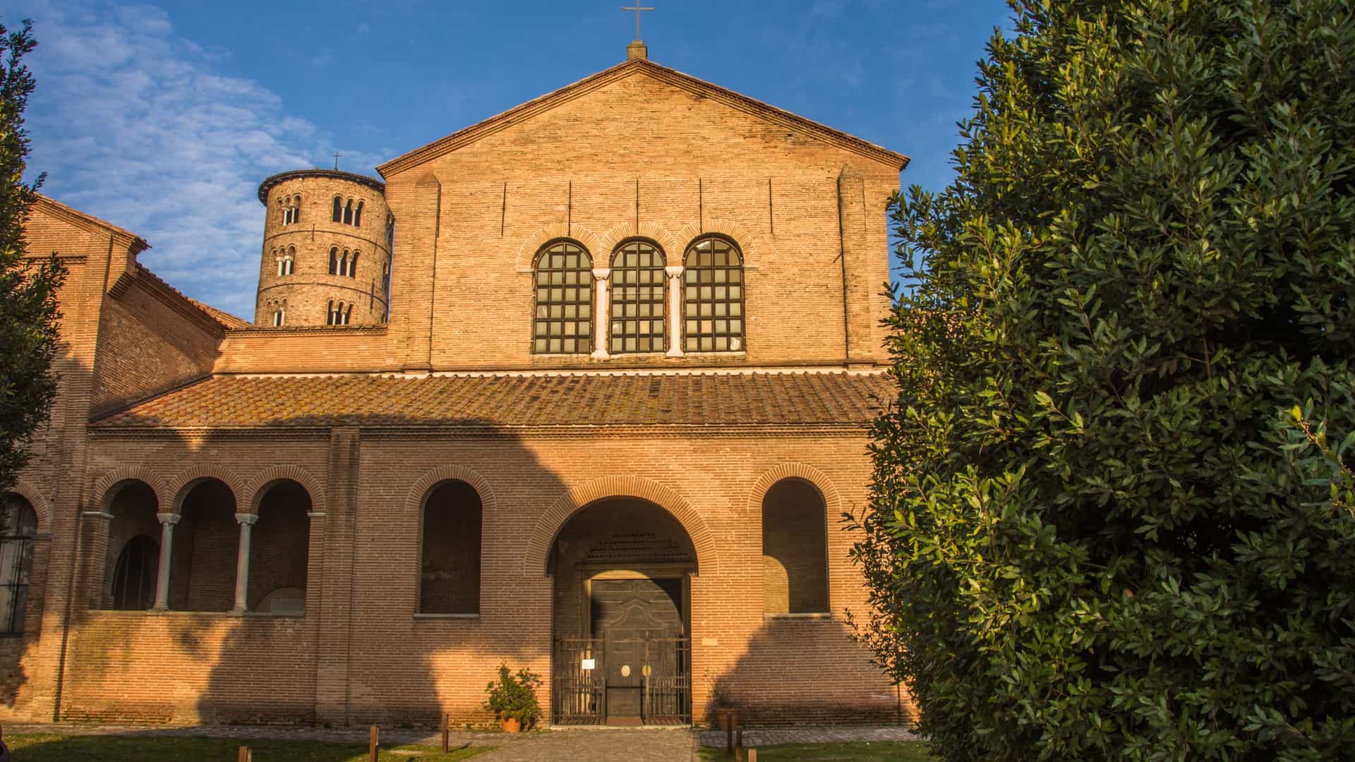 The exterior view of the Basilica of Sant'Apollinare in Classe, a 6th century Early Christian church and UNESCO World Heritage site in Ravenna, Italy. The photo shows the simple, imposing facade made of red brick, with an attached rectangular narthex and the tall, cylindrical, nine story bell tower (campanile) on the right side. The basilica is set in a wide, flat, grassy field under a bright sky.