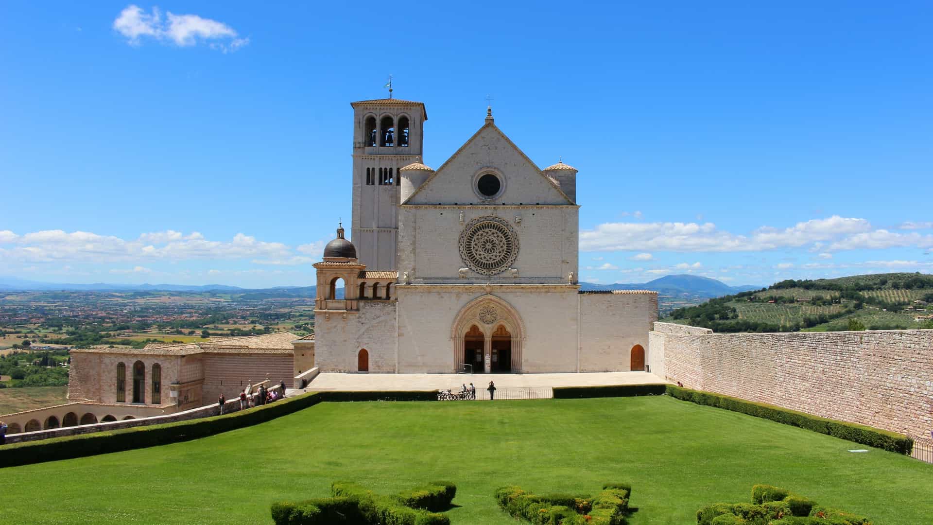 A scenic, elevated view of the Basilica di San Francesco (Basilica of Saint Francis) in Assisi, Italy. The complex, consisting of the two tiered church and the Sacro Convento (Sacred Convent), is built with white and pink stone and sits on the edge of the hill town. It overlooks a vast, green Umbrian valley with a patchwork of cultivated fields extending to the horizon.