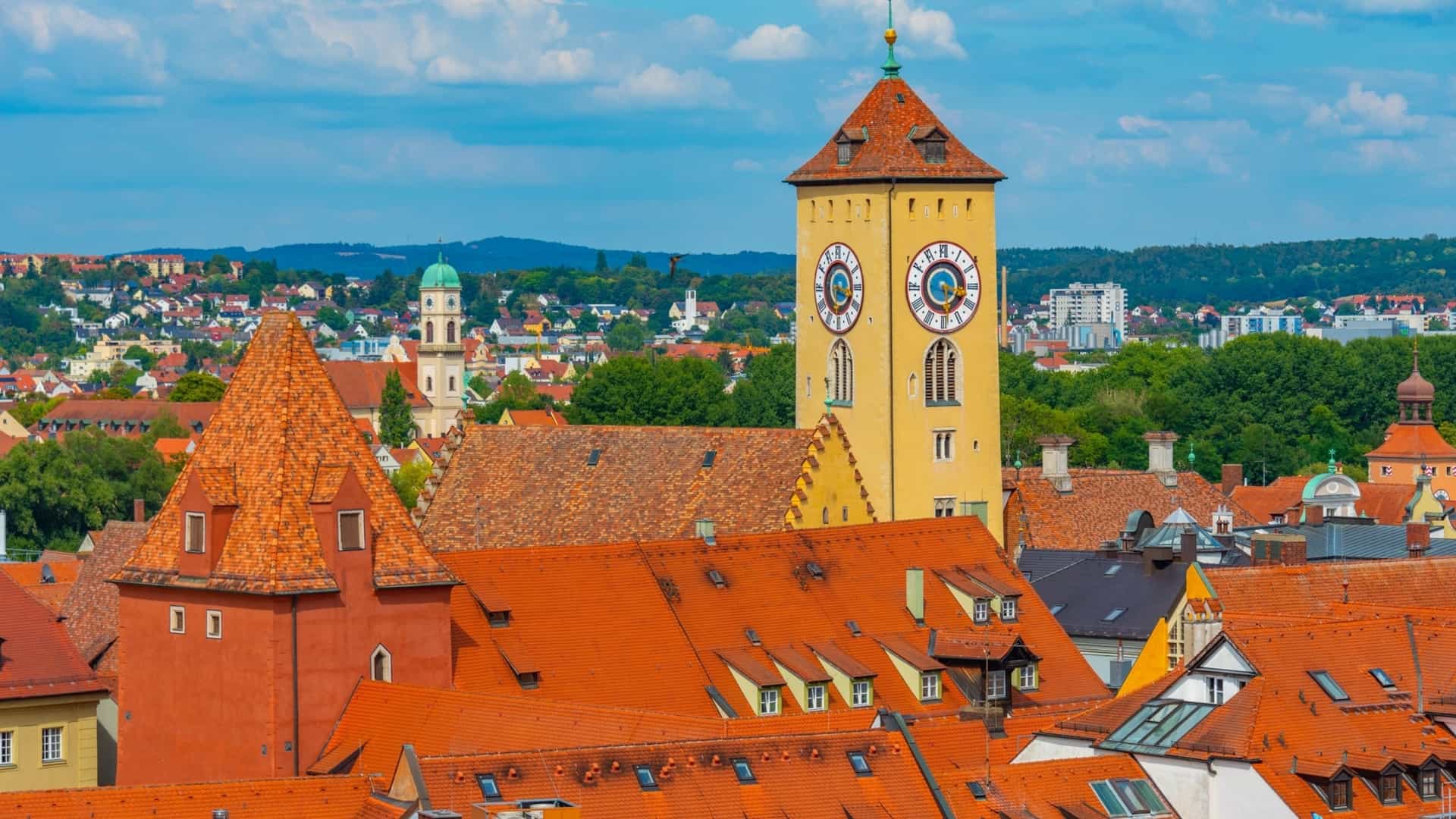 The image captures the Town Hall's unique location, its highly decorative, frescoed facade with trompe l'oeil painting, and the surrounding historic cityscape of half timbered and tiled roof buildings along the riverbanks. The river flows around the structure, connecting the two parts of the city.