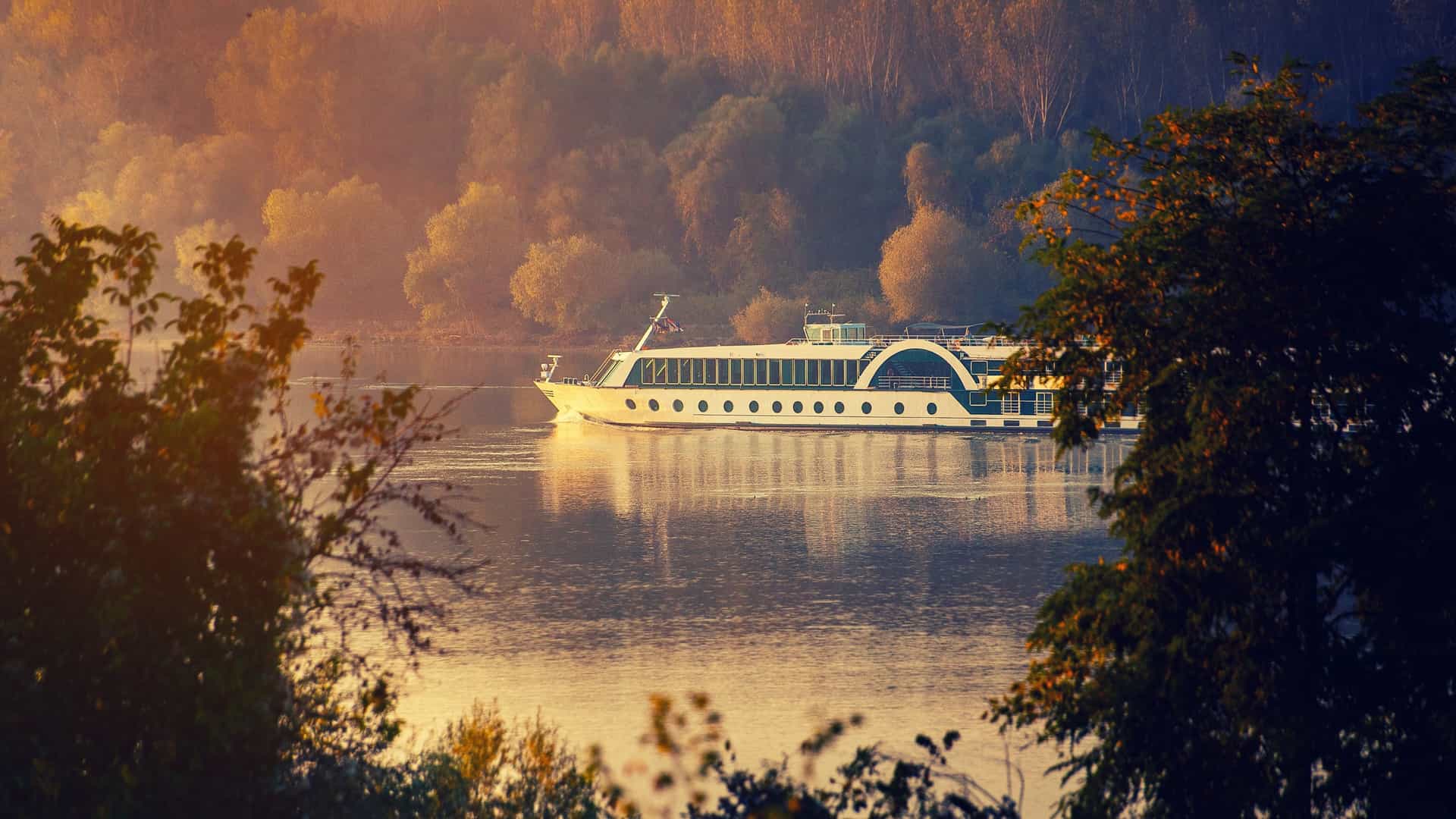 A long, white river cruise ship sails along the Danube River, passing historic central European buildings and forested hills under a blue sky.