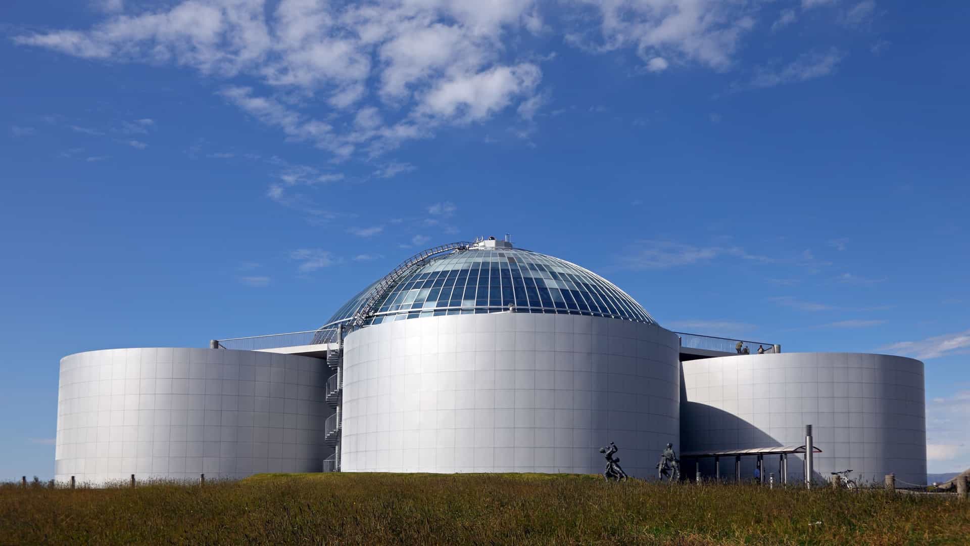 A sunny exterior view of the Perlan (The Pearl) museum and observation deck in Reykjavik, Iceland. The modern structure consists of a large glass dome surrounded by several huge, silvery cylindrical geothermal hot water storage tanks, set atop a grassy hill against a bright blue sky.