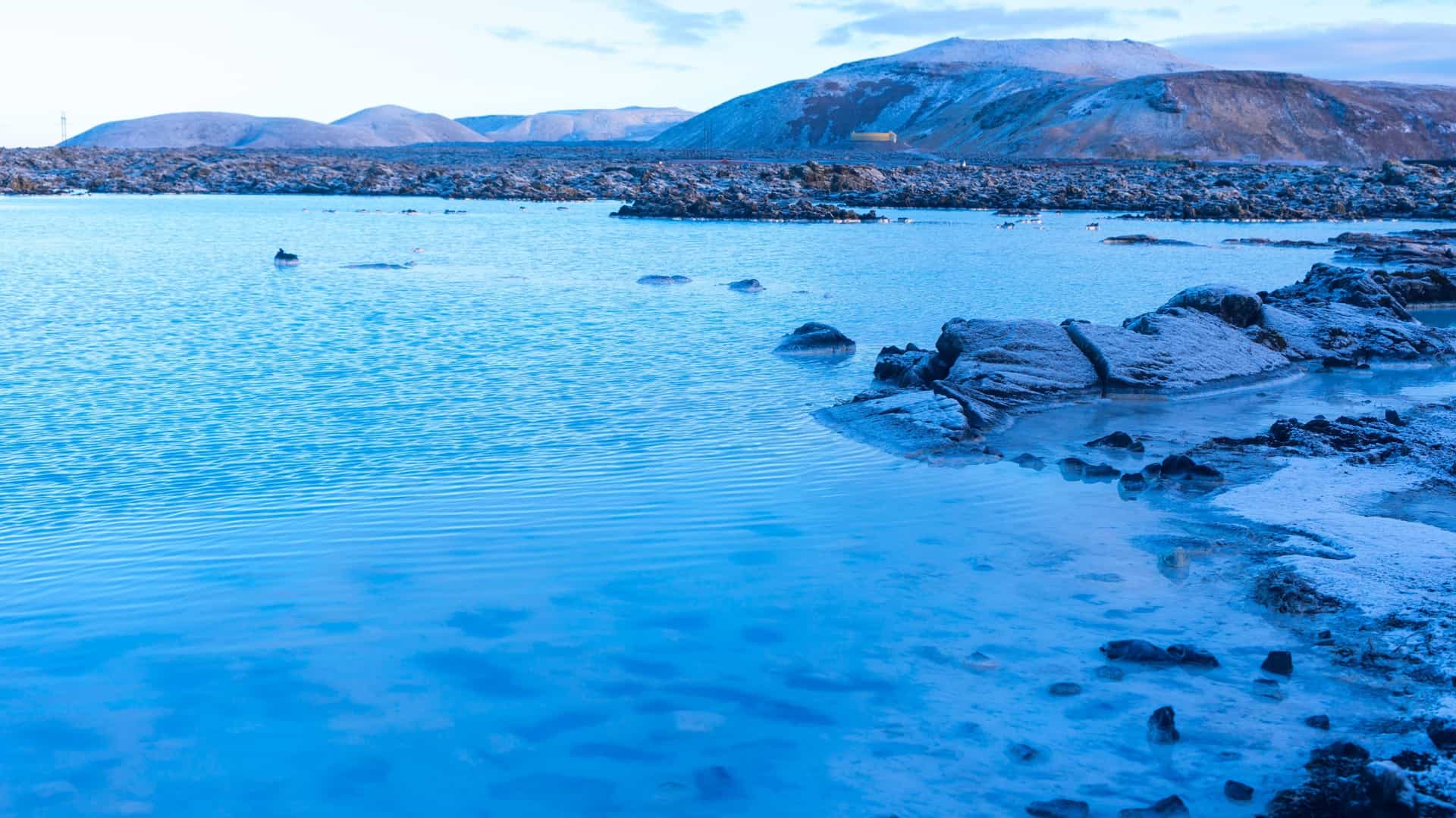 A serene, milky blue geothermal spa known as the Blue Lagoon in Iceland. The warm, mineral rich water, dense with silica, is surrounded by rough, black lava rocks partially covered in green moss, with steam rising from the surface into the clear sky.