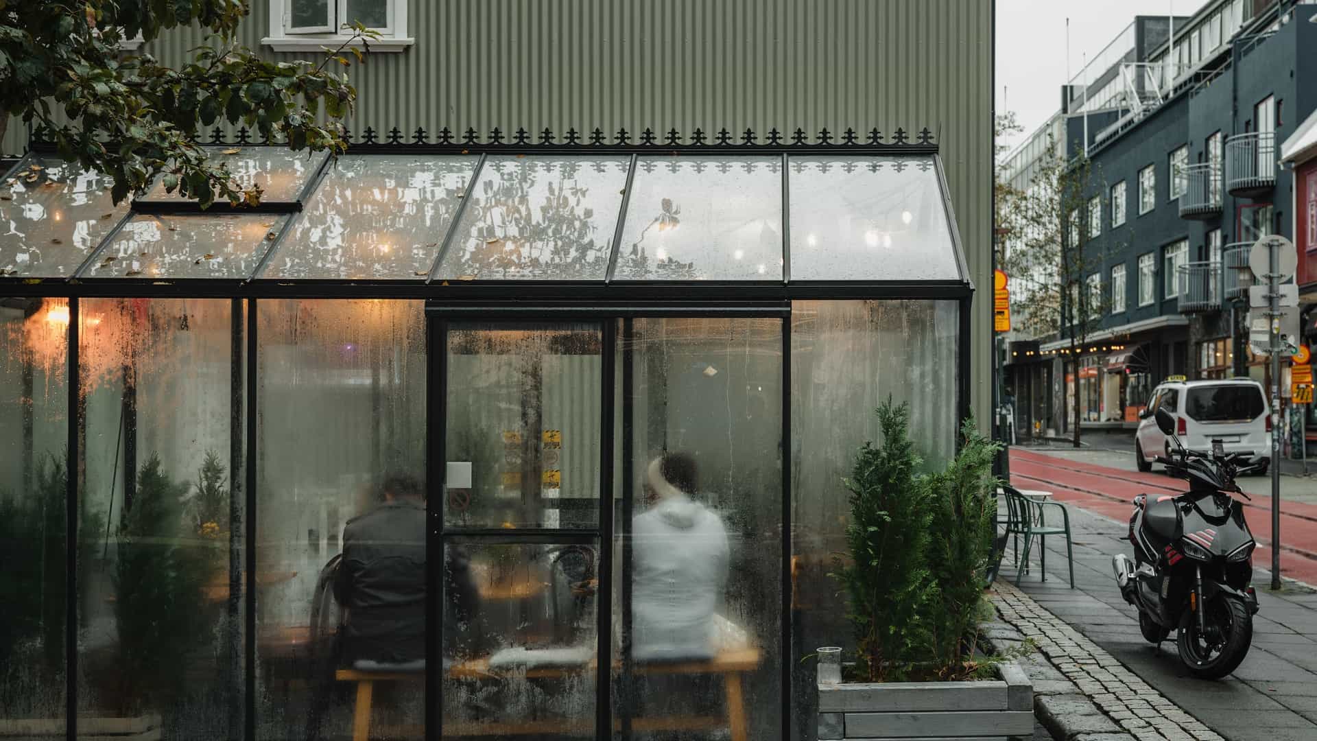 A winter scene of a cozy street side cafe, most likely on Laugavegur, the famous shopping street in Reykjavík, Iceland. The cafe's large window is visible, with a small seating area outside and a clear view of the street, which is covered in a light dusting of snow. The surrounding buildings are in the traditional, colorful Icelandic style.