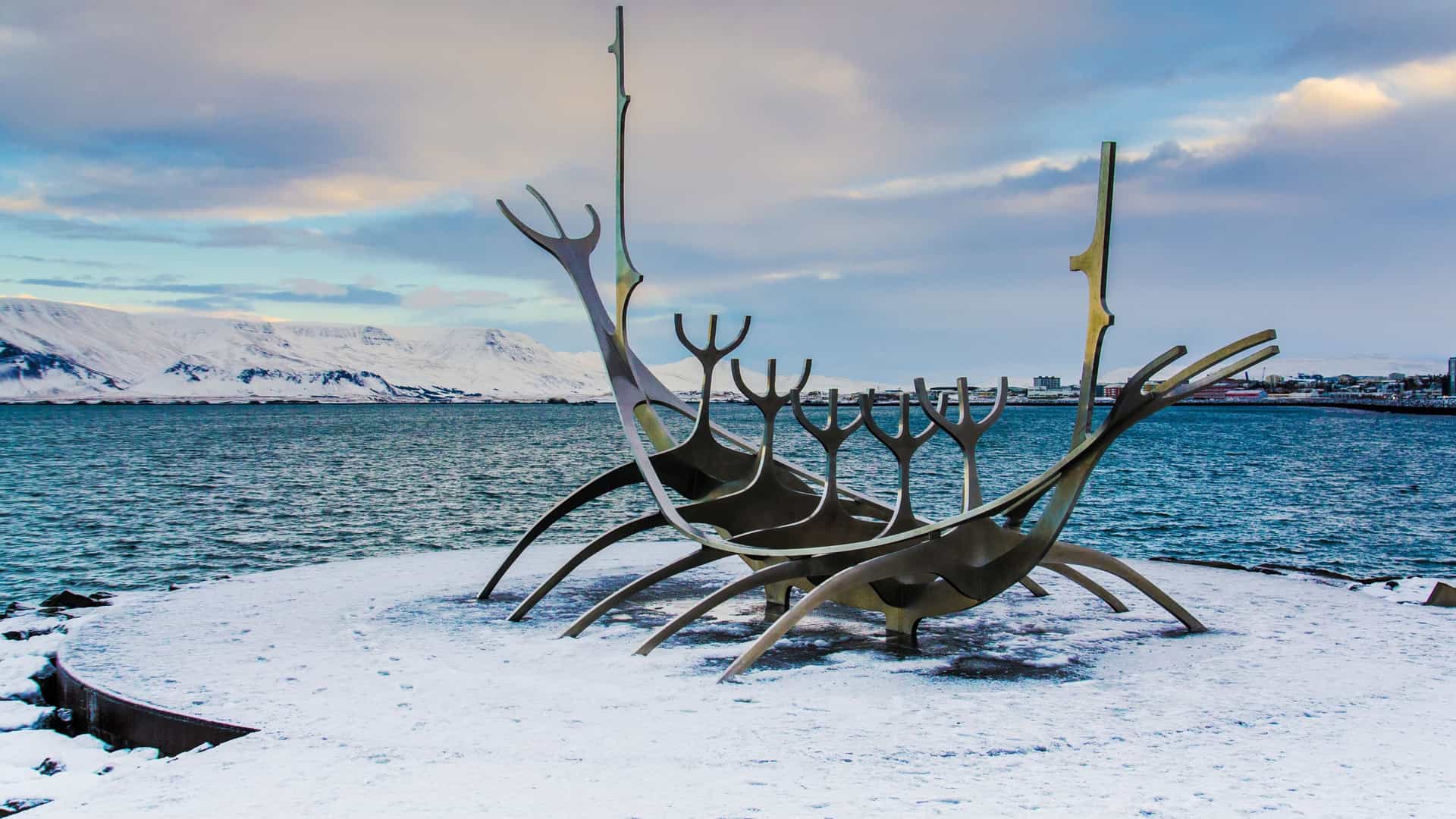 The 'Sun Voyager' (Sólfar), a striking stainless steel sculpture resembling a skeletal Viking longship, stands on a granite platform on the seaside in Reykjavík, Iceland. The sculpture points out towards the ocean and the silhouette of Mount Esja in the distance, under a bright sky.