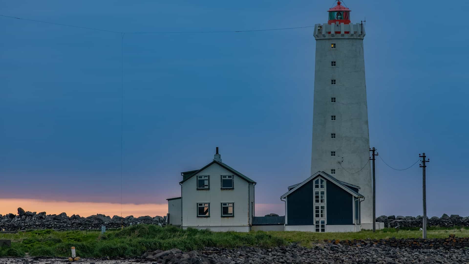 A dramatic view of the Grótta Lighthouse in Seltjarnarnes, Iceland, a striking white structure standing on a small tidal island connected to the mainland by a narrow causeway. The scene features dark, rugged shoreline rocks and the vast, overcast Atlantic Ocean.