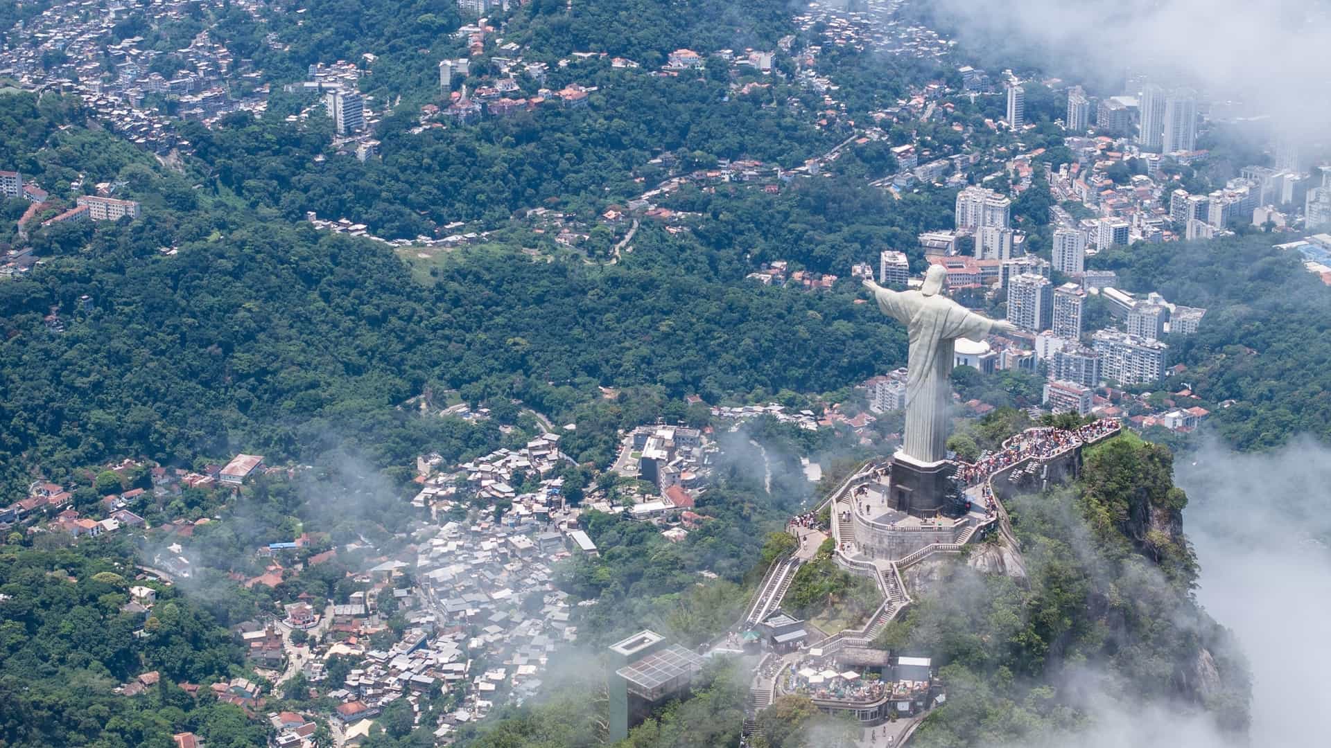An aerial view of the Christ the Redeemer statue on Corcovado mountain, overlooking the sprawling city of Rio de Janeiro, Brazil. The statue with its outstretched arms dominates the peak, while the background shows a vast urban area nestled between green mountains and the sparkling blue of Guanabara Bay and the Atlantic Ocean.