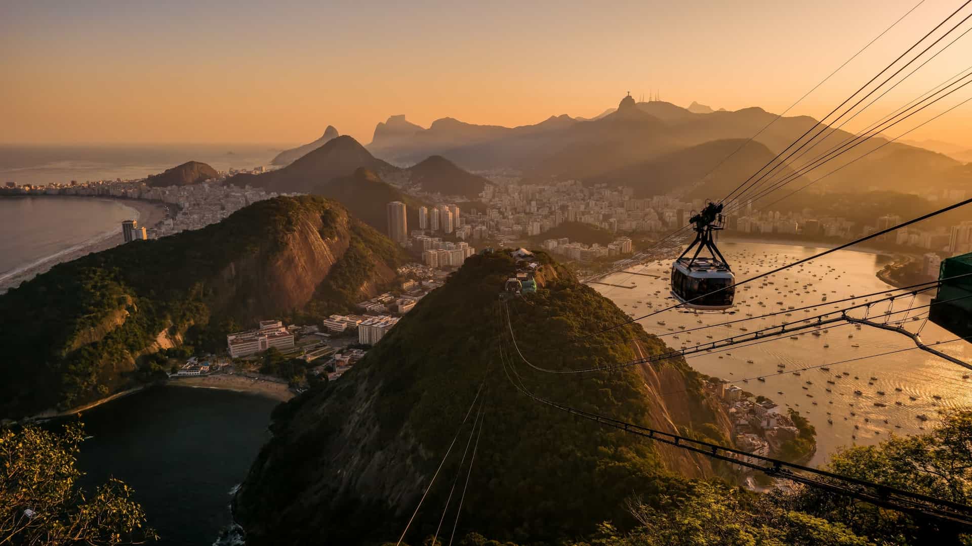 A photograph of the Sugarloaf Mountain cable car (Bondinho do Pão de Açúcar) ascending its steep cable line, with a breathtaking panoramic view of Rio de Janeiro in the background. The scene includes the sweeping coastline of Guanabara Bay, the city's urban sprawl, and the distant mountains, all under a clear sky. A cable car cabin is visible near the center, suspended above the green hills and blue water.