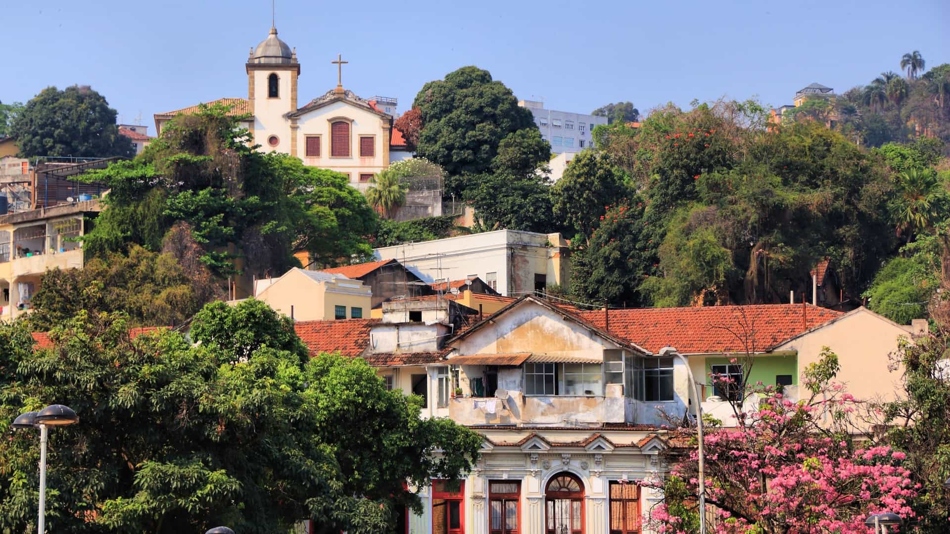 A view of a steep, winding cobblestone street in the Santa Teresa neighborhood of Rio de Janeiro, lined with old colonial style buildings and lush tropical greenery. The image is characteristic of the area's bohemian and historic charm.
