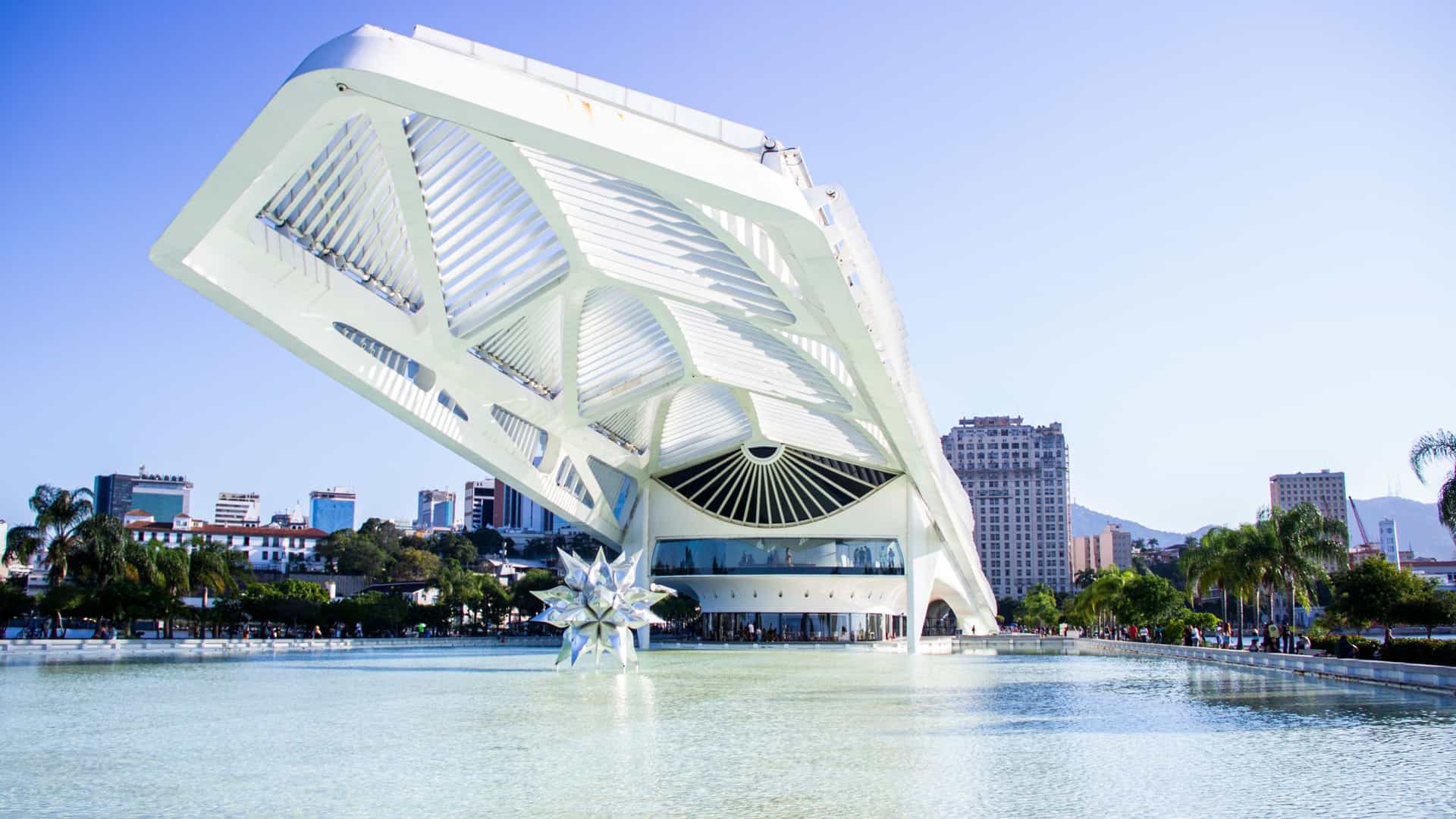The exterior of the Museu do Amanhã (Museum of Tomorrow) in Rio de Janeiro. The white, futuristic, neofuturistic building, designed by Santiago Calatrava, features a long, skeletal, wing like roof structure extending over a large reflecting pool on the waterfront of Guanabara Bay.