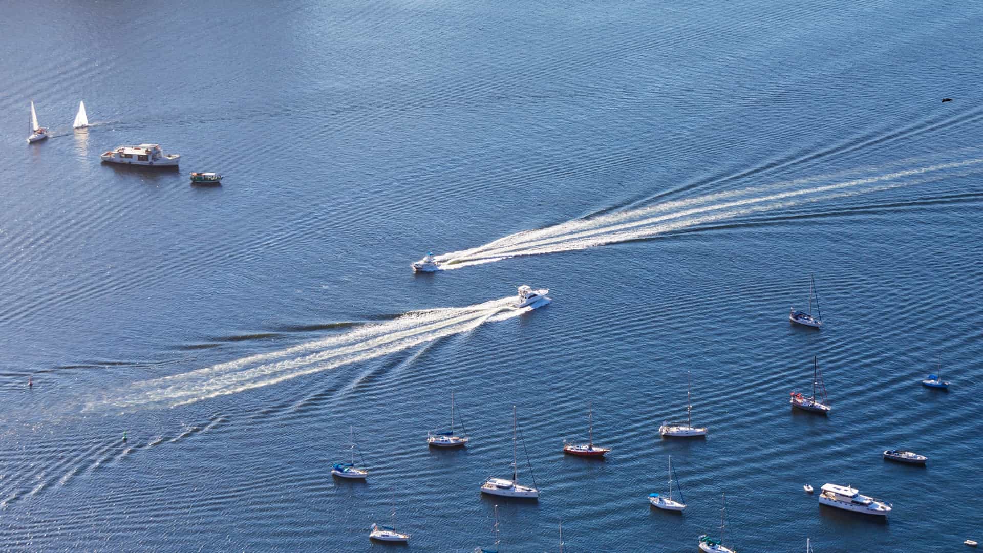A bright, sunlit view of Guanabara Bay, with several sailboats and yachts scattered across the water in the foreground. In the background, the iconic, rounded granite peaks of Sugarloaf Mountain (Pão de Açúcar) and Urca Hill (Morro da Urca) dominate the horizon, with the city's coastal buildings visible along the shore.