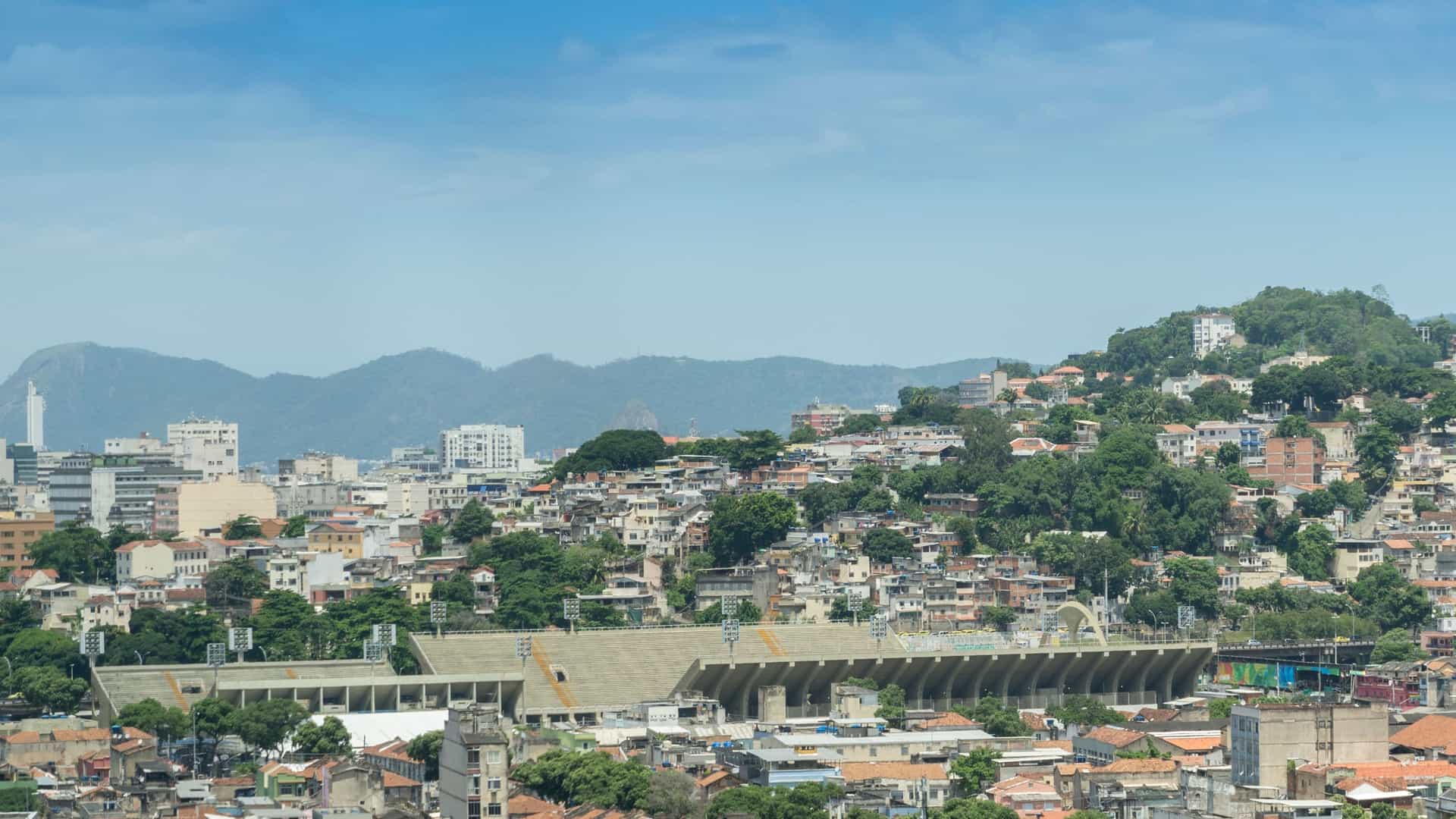 An aerial or long distance view of the Sambadrome Marquês de Sapucaí in Rio de Janeiro, a purpose built, straight parade avenue lined on both sides by white concrete grandstands for spectators. The avenue culminates at the iconic, monumental arch of the Apotheosis Square at the far end.