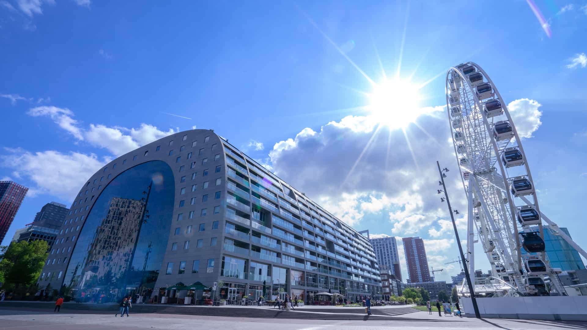 A modern, horseshoe shaped building in Rotterdam called the Markthal. The grey stone structure functions as both a residential/office complex and a covered market hall, featuring a vast, segmented glass cable facade on its end, which reflects the surrounding urban scene.