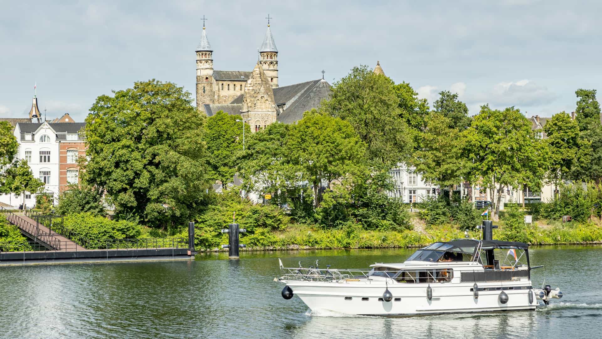 A boat (likely a small vessel) sails across the calm waters of the Maas River (Nieuwe Maas) in Rotterdam, Netherlands. The tranquil river surface reflects the sky. In the background, the dense, modern cityscape of Rotterdam's high rise buildings and potentially landmarks like the Erasmus Bridge define the horizon.