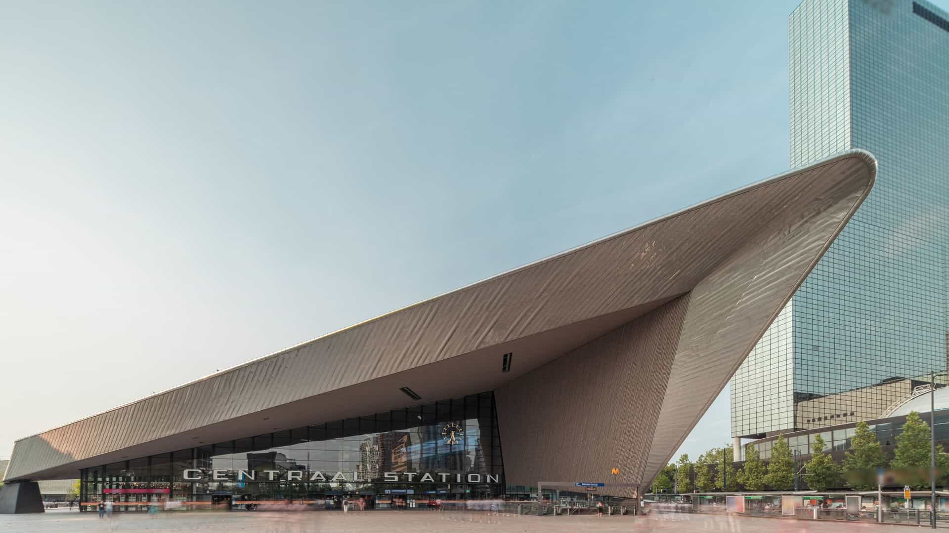A hyperlapse shot of the modern Rotterdam Central Station, featuring its large, distinctive, angular main entrance and the bustling plaza in front, with the background dominated by the city's modern skyscrapers.