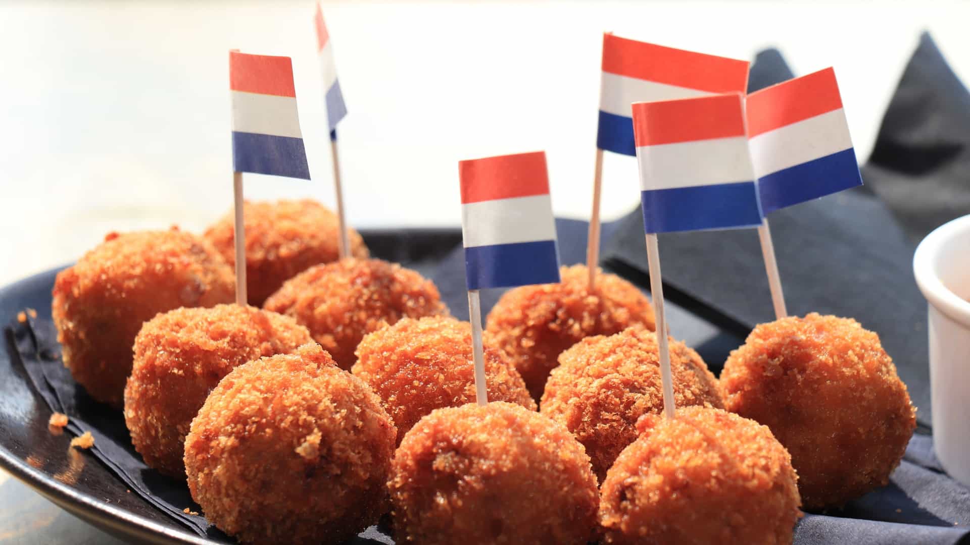 A close up image of several piping hot, golden brown Dutch bitterballen (deep fried savory meatballs or croquettes) served as a classic snack, typically presented on a plate next to a small cup or dollop of sharp yellow mustard for dipping.