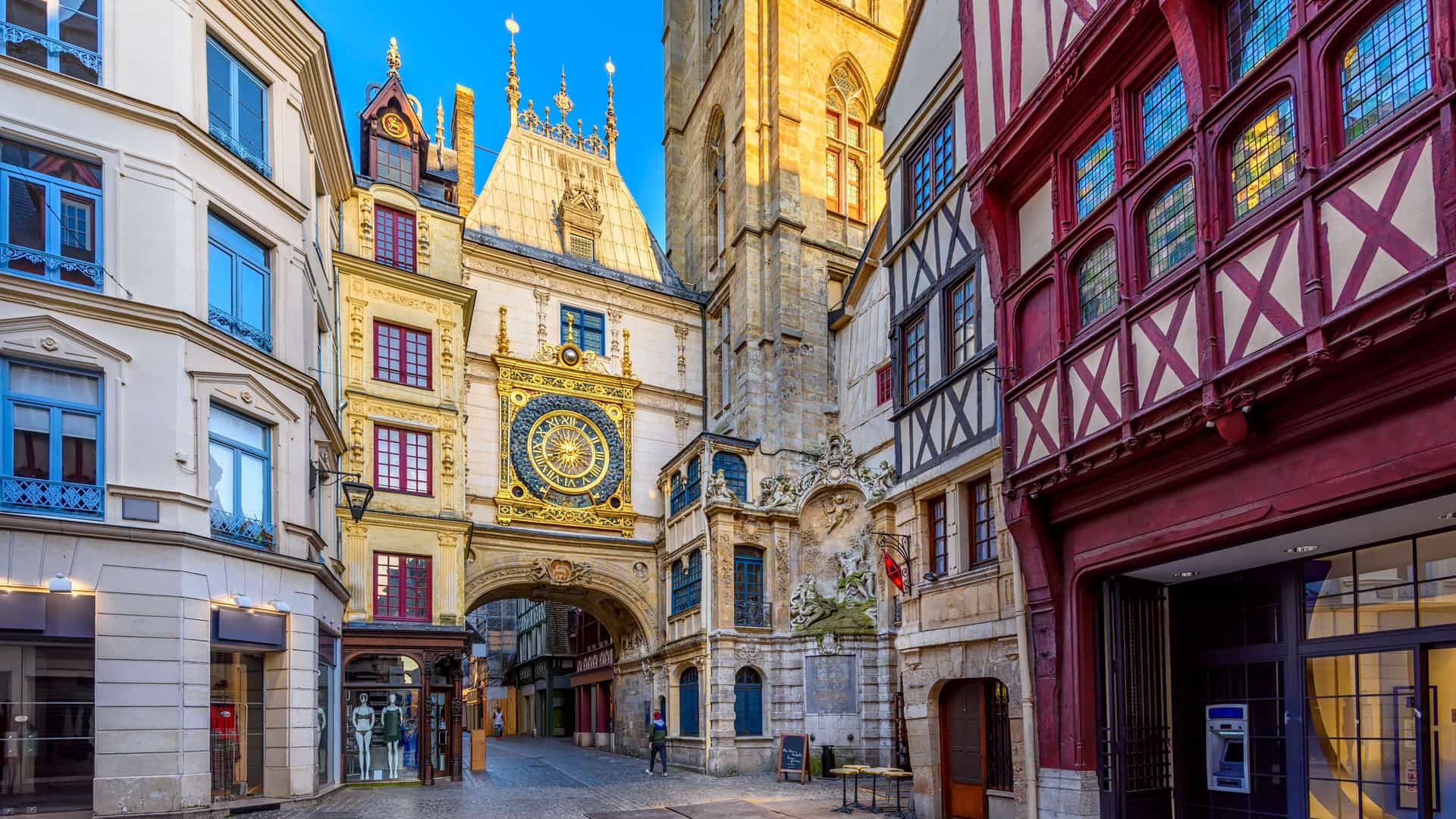 A view of the Gros Horloge (Great Clock) in Rouen, France, a 14th century astronomical clock set within a Renaissance archway that spans a pedestrian street. The golden clock face displays 24 sun rays against a starry blue background, featuring a single hour hand tipped with a lamb. The arch is attached to a tall Gothic belfry (bell tower), and historic half timbered houses line the cobblestone street.