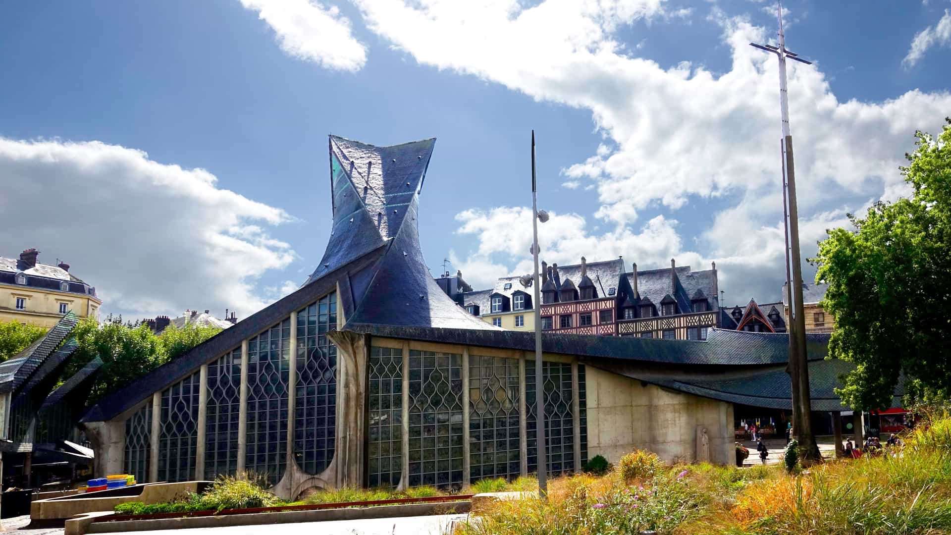 A close-up view of the distinctive, modern architecture of the Church of Saint Joan of Arc (Église Sainte-Jeanne-d'Arc) in Rouen, France, showing the dramatic, dark, scale-like roof curving like flames or an overturned boat above a large, transparent glass facade in the historic Old Market Square.