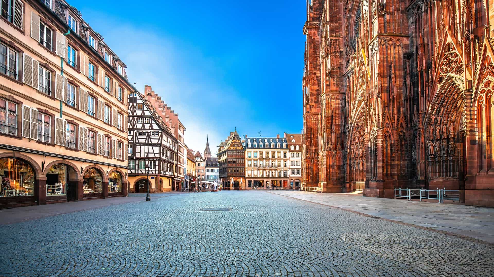 An iconic view of the Place de la Cathédrale in Strasbourg, France, featuring the intricate Gothic façade and single, towering spire of the Notre-Dame de Strasbourg Cathedral. The square is bustling with activity and surrounded by historic half-timbered buildings.