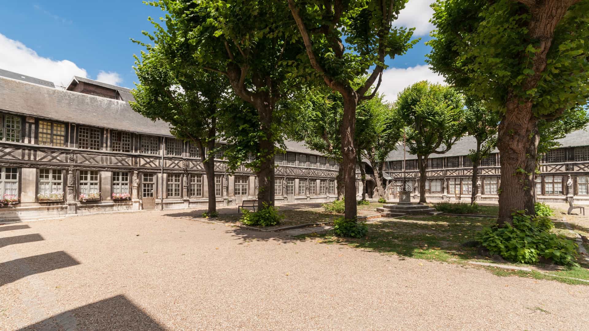 A wide-angle view of the historic Aître Saint-Maclou courtyard in Rouen, France, showcasing the inner quadrangle surrounded by preserved 16th-century half-timbered buildings. The ground is covered in light gravel, and several large, leafy trees dominate the frame under a bright, clear blue sky.