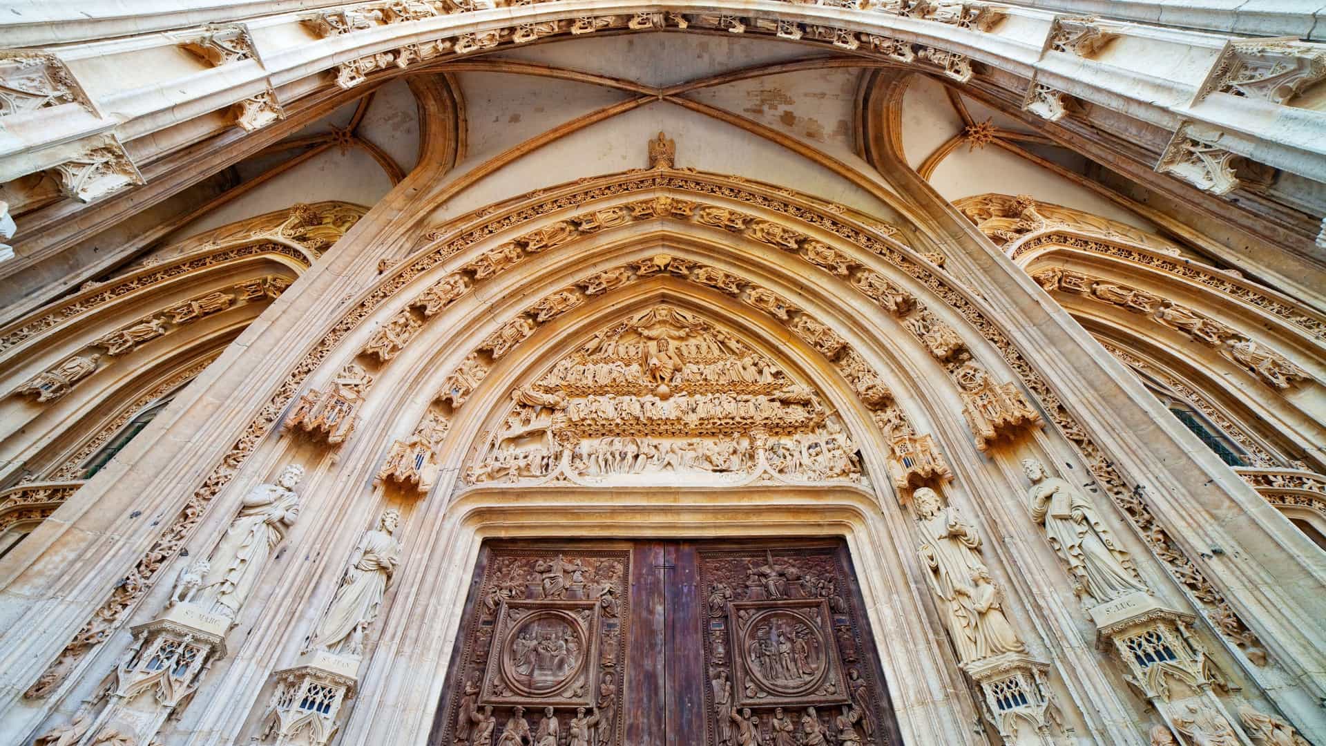 close up view of the magnificent pentagonal porch of the Church of Saint Maclou in Rouen, France, a masterpiece of Flamboyant Gothic architecture. The porch features five gabled arches arranged in a semicircle, adorned with intricate, flame like stone tracery. The three central bays hold the church's Renaissance carved wooden doors, and the tympanum of the main portal is sculpted with a depiction of the Last Judgement.