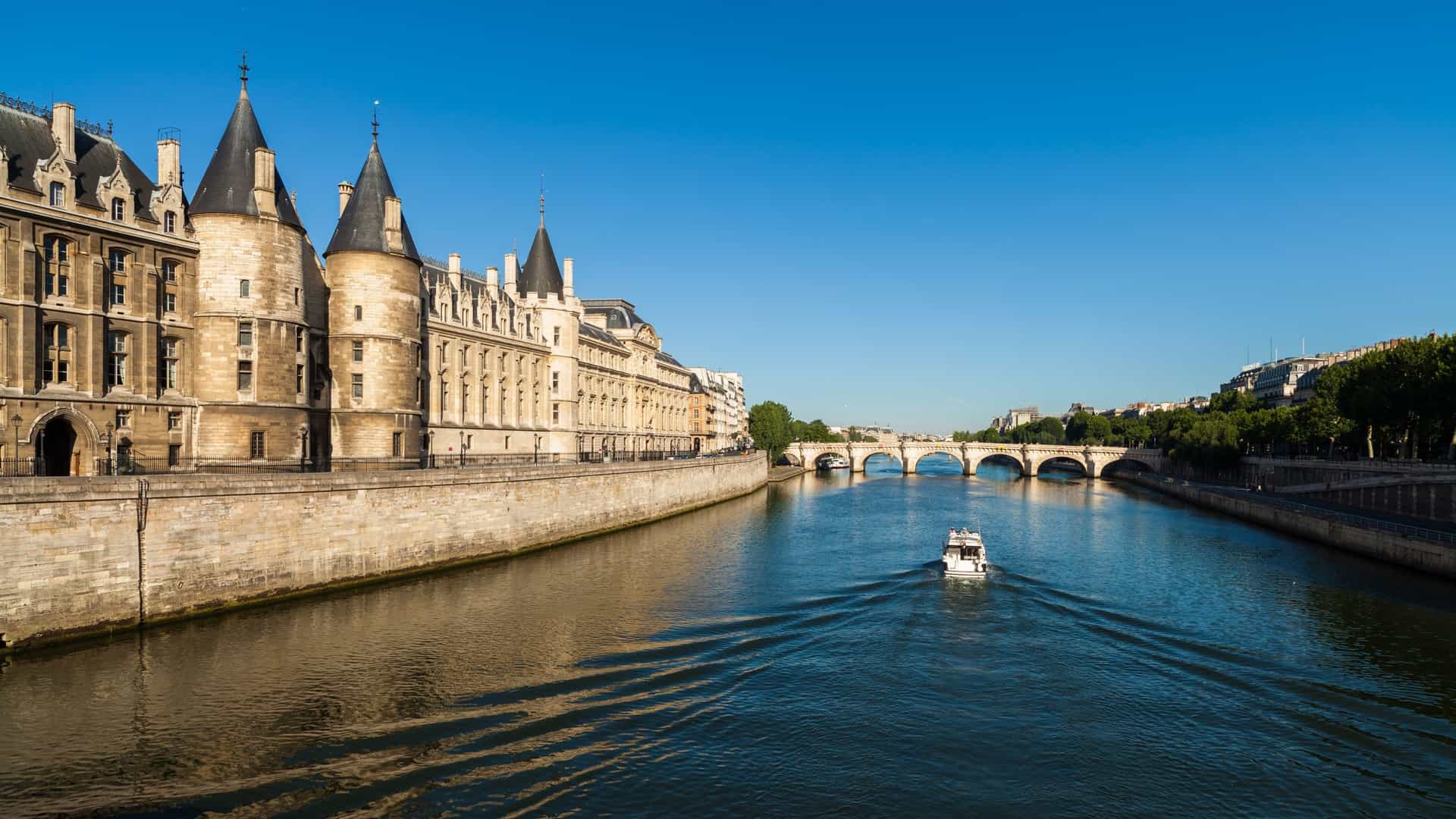 A scenic photograph of the River Seine in Paris, France, likely taken around sunset or twilight. A tourist cruise boat, such as a Bateau Mouche, with large windows and multiple decks, is sailing on the water. The surrounding riverbanks are lined with iconic Parisian architecture, featuring historical stone buildings and bridges that are beginning to light up as the day ends.