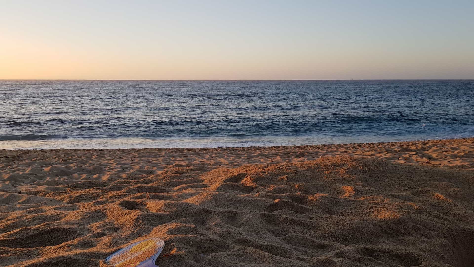 A serene sunset over El Quisco beach in Chile. The sun is setting over the Pacific Ocean, casting a brilliant orange and red glow on the sky and reflecting on the water, with silhouetted coastal rock formations or cliffs visible in the foreground.