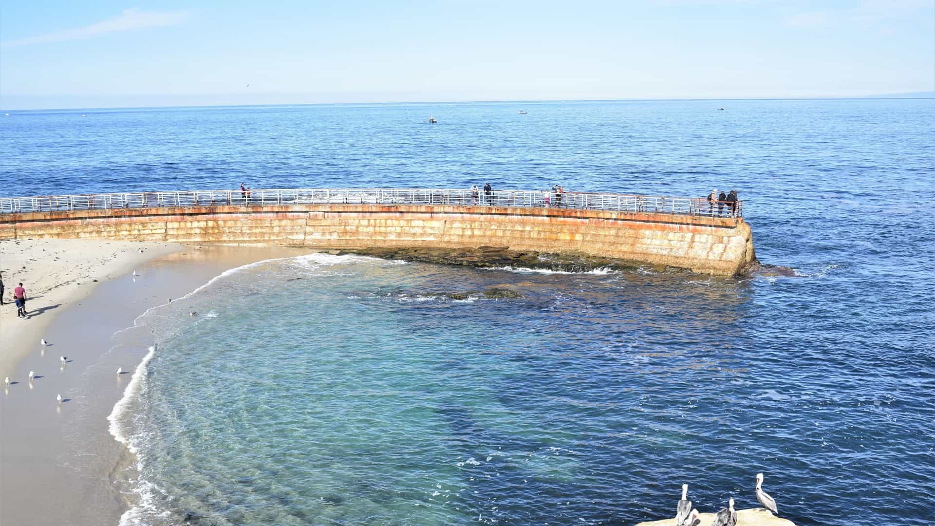 A scenic view of La Jolla Cove in San Diego, California, featuring a small, sandy beach nestled between rugged sandstone cliffs and the bright, turquoise water of the Pacific Ocean.