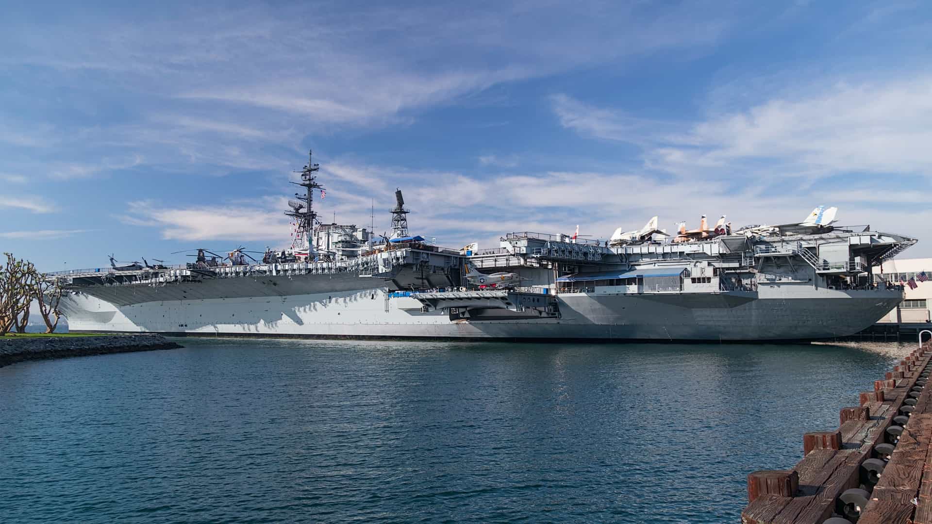 A large, exterior view of the historic USS Midway aircraft carrier, now the USS Midway Museum, docked at Navy Pier in San Diego, California. The massive gray ship, featuring its flight deck and control tower (island), dominates the waterfront.