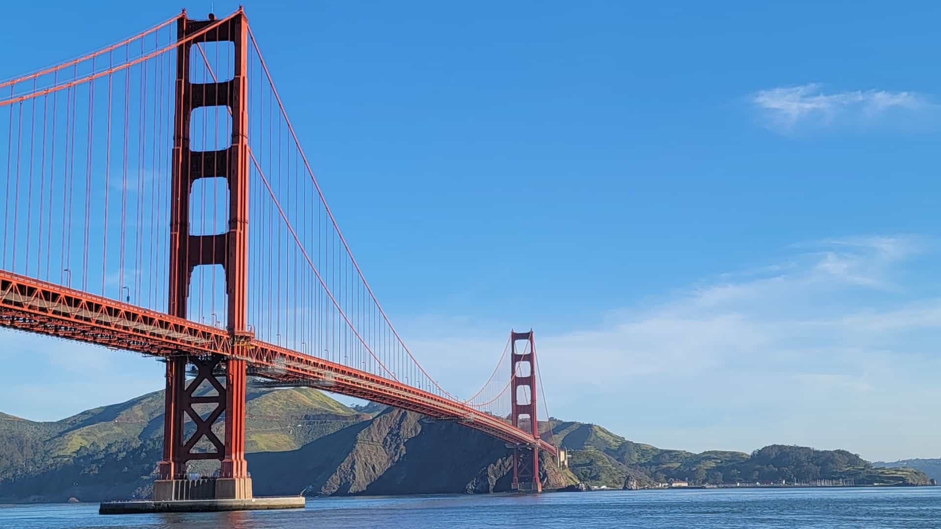 A bright, clear day view of the iconic Golden Gate Bridge in San Francisco, California. The famous orange red suspension bridge stretches across the water, with its towers highlighted against a vivid blue sky that has some scattered white clouds. The water below is calm, and the hills of the Marin Headlands are visible in the distance.