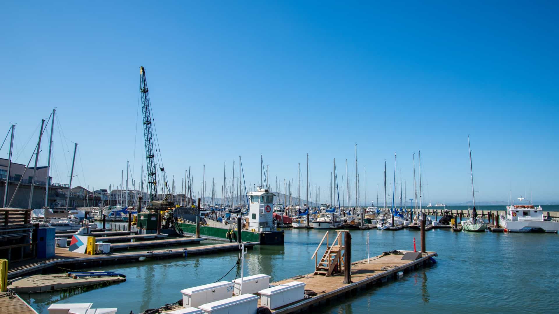 A bright, bustling scene at Fisherman's Wharf in San Francisco, California, featuring a crowded street lined with colorful shops, seafood stalls, and restaurant signs. In the background, the San Francisco Bay is visible with moored fishing boats and a partial view of a nearby pier.