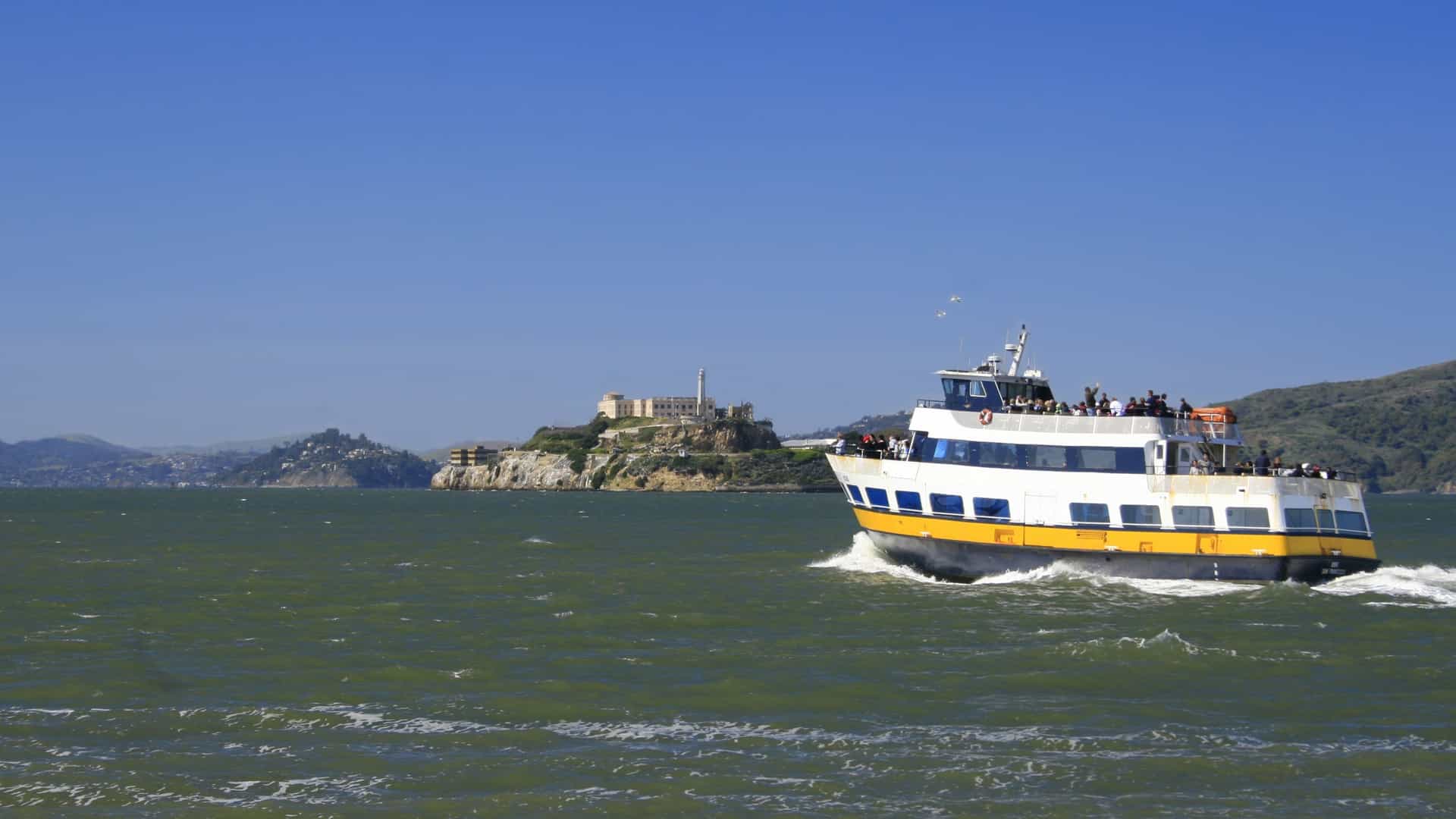 View of a large red and white tourist boat cruising past Alcatraz Island in the choppy waters of the San Francisco Bay, with the former prison buildings visible on the rocky island.