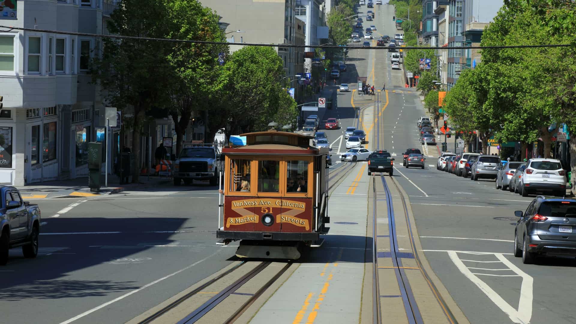 A classic, compressed view of a double ended San Francisco cable car (likely maroon colored) ascending or descending the steep incline of California Street. The street extends dramatically in the background, lined by tall downtown buildings and skyscrapers, capturing the iconic urban landscape of San Francisco.