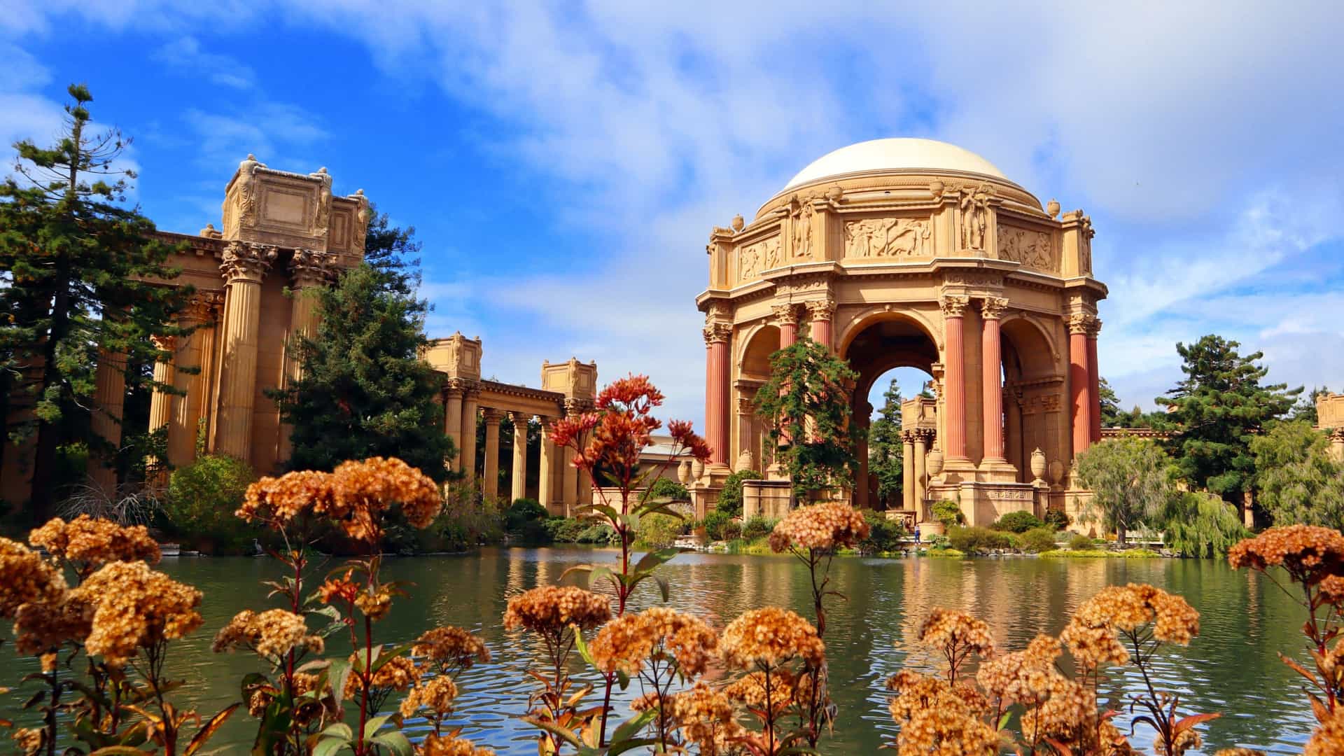 A photograph of the iconic Palace of Fine Arts in San Francisco, California. The image features the grand, Greco Roman style rotunda and a wide, curved colonnade reflected in the tranquil lagoon, with greenery surrounding the structure. The architecture is designed to evoke the romantic impression of an ancient Roman ruin.