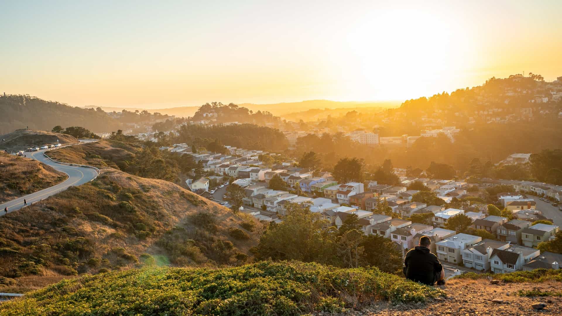 A beautiful photograph of a warm sunset over the city of San Francisco, taken from the vantage point of Twin Peaks. The view captures the city's skyline and surrounding urban area bathed in the golden orange light of the setting sun, which creates a serene and picturesque scene.