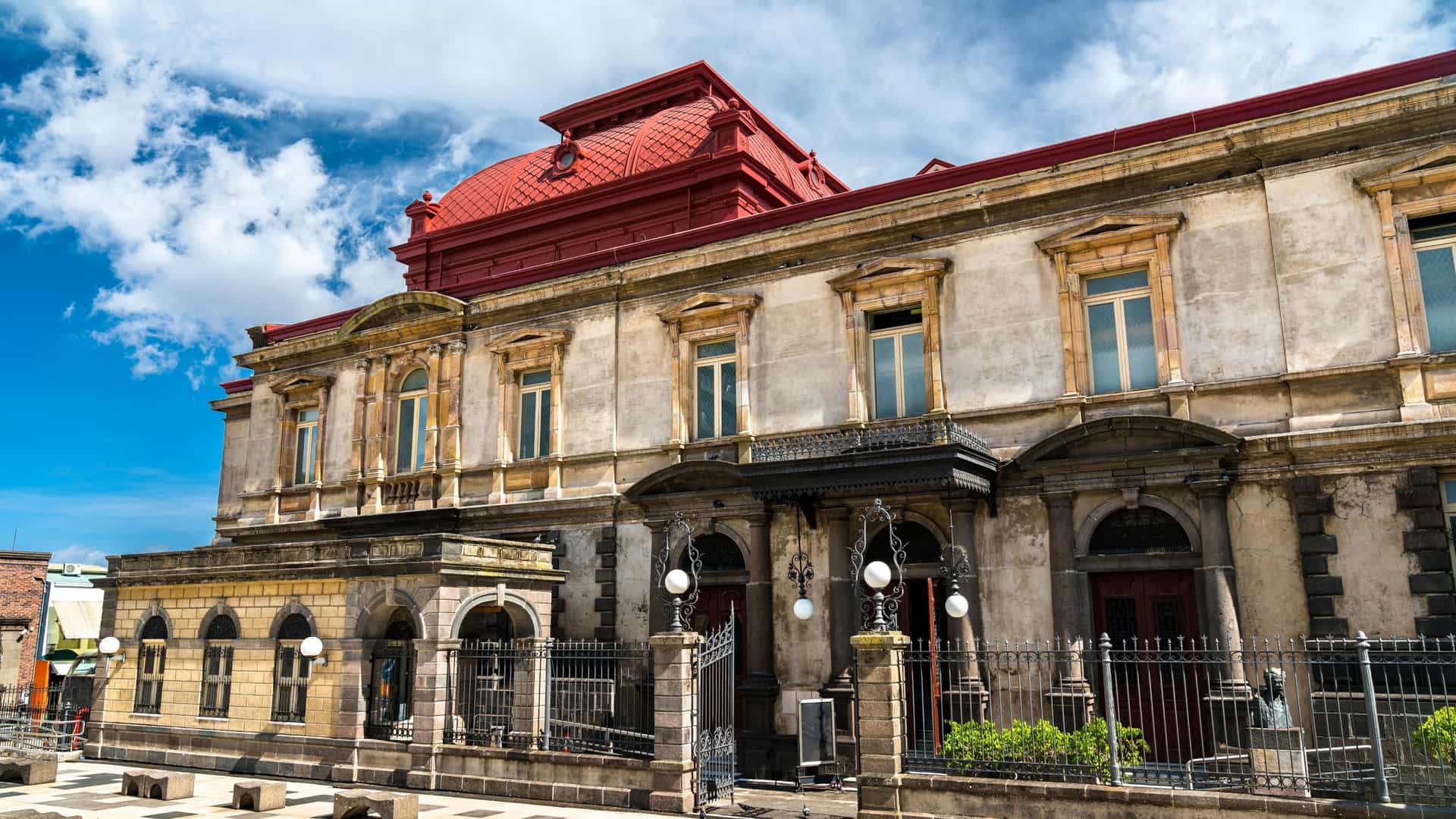 The classical architectural facade of the National Theatre of Costa Rica in San José, featuring a prominent entrance with columns and statues on the upper story, all set against a clear blue sky.