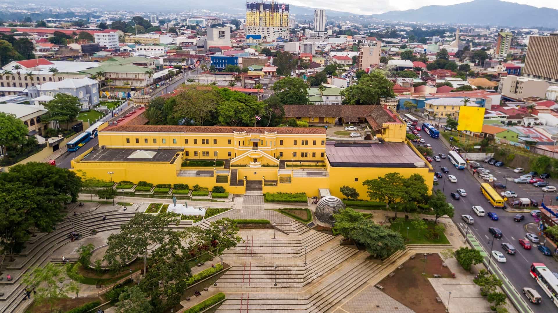 A beautiful aerial view of a prominent yellow building, identified as the National Museum of Costa Rica (Museo Nacional de Costa Rica), surrounded by the urban landscape of San José.