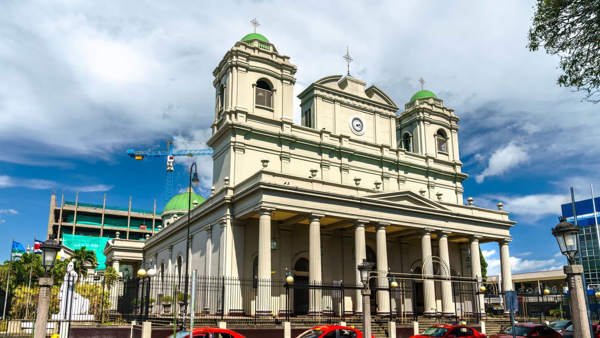 The Metropolitan Cathedral of San José, Costa Rica, displaying its neoclassical and Renaissance style architecture. The front facade, featuring a central pediment and bell towers, is framed by lush tropical trees, with the adjacent Parque Central visible in the foreground.