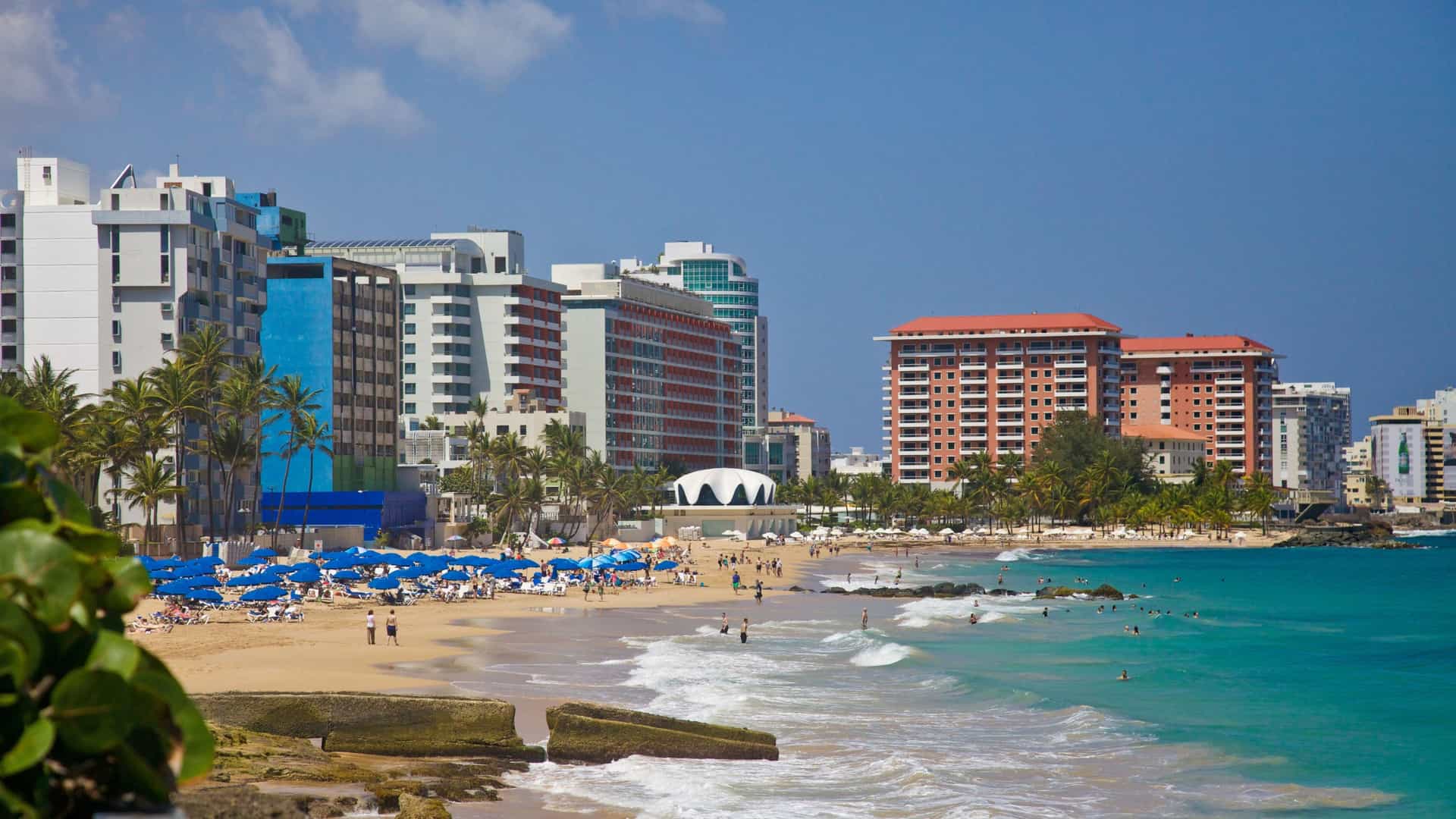 A scenic view of Condado Beach in San Juan, Puerto Rico, featuring golden sand, turquoise Atlantic ocean waves, and the urban backdrop of high rise resort hotels and residential buildings.