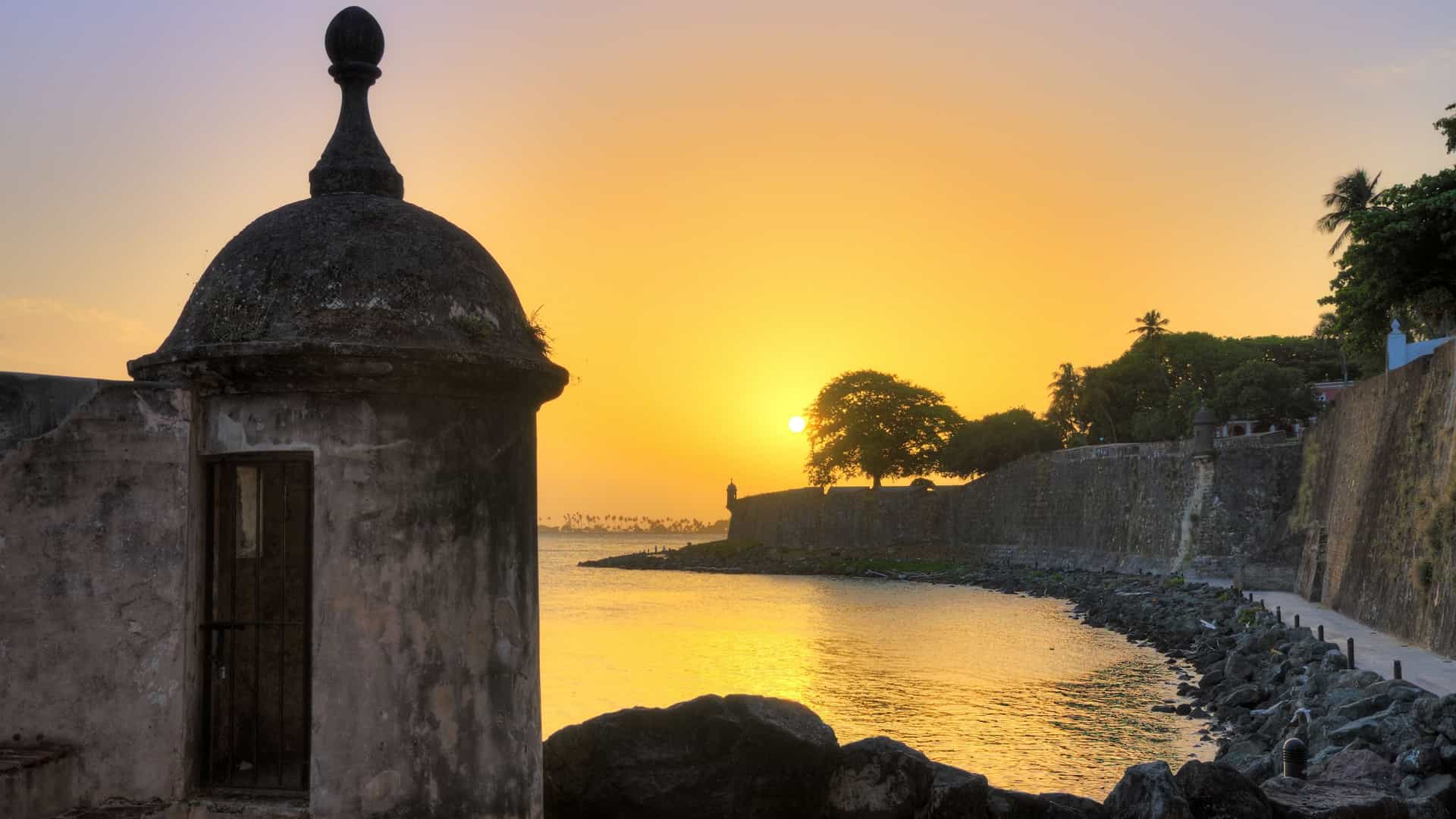 A beautiful summer sunset view of the outer wall and sentry box (garita) of Castillo San Felipe del Morro in Old San Juan, Puerto Rico. The stone walls of the Spanish colonial fort are silhouetted against a colorful evening sky over the ocean.