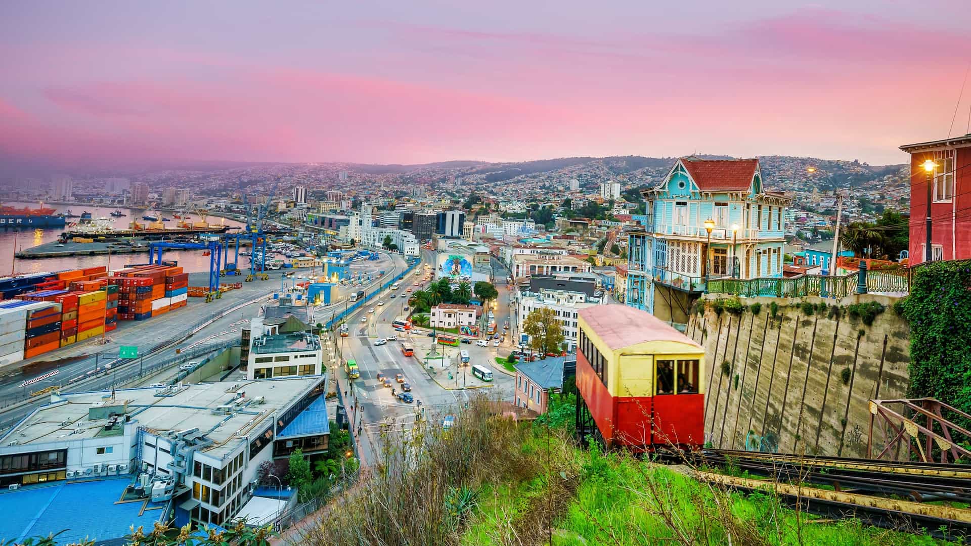 A passenger carriage of a historic funicular railway (ascensor) in Valparaíso, Chile, moves along its steep tracks. The carriage is set against the densely built hill, featuring a patchwork of colorful, urban houses, characteristic of this UNESCO World Heritage city.